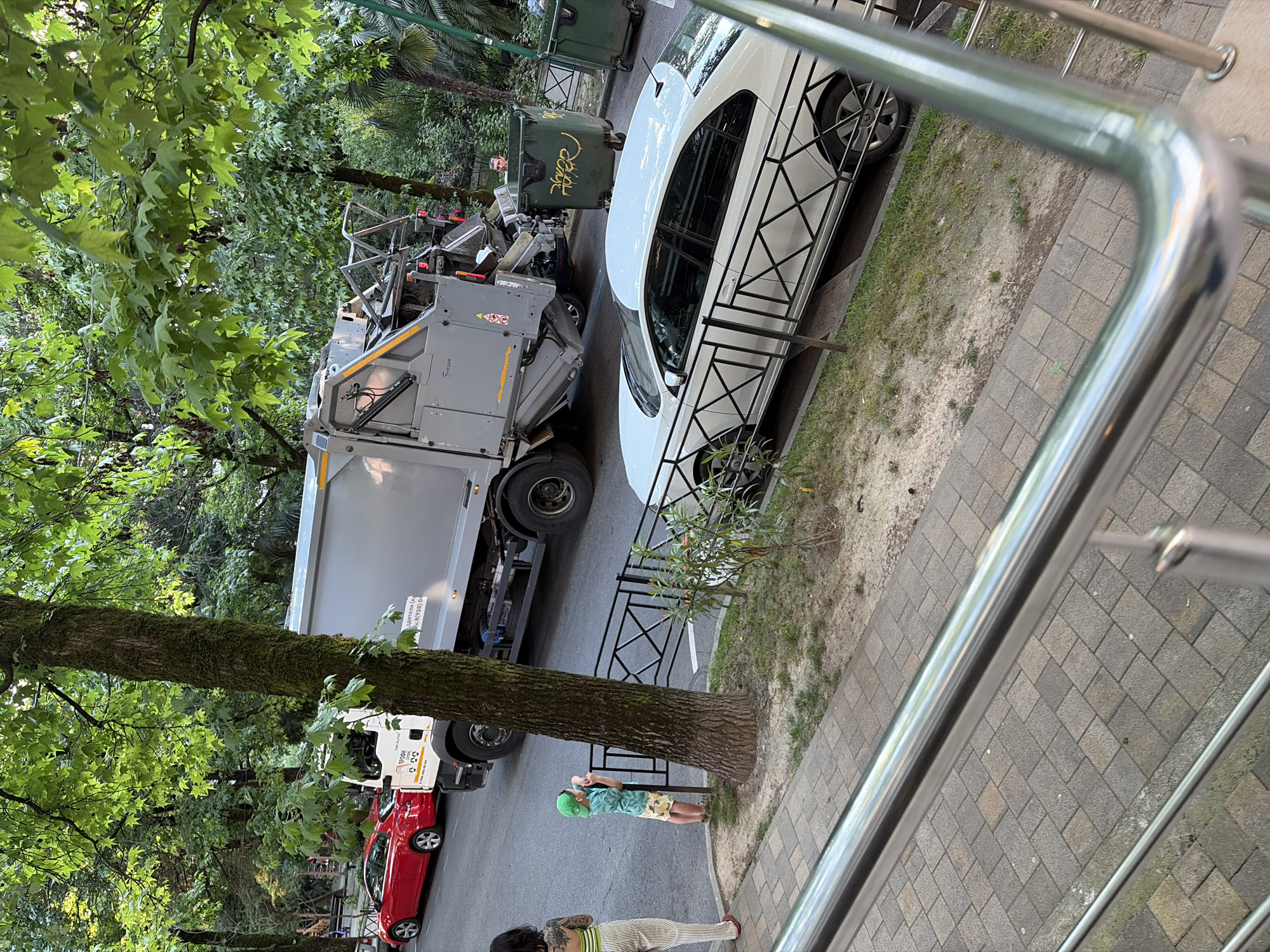 A garbage truck is parked on a street in Sochi, Russia, next to a white car. The garbage truck is gray with a large compartment for waste. The white car has a distinctive black geometric pattern on its side. In the background, there are green trees and some buildings. A person wearing a green hat and a patterned shirt is standing near a trash bin. Another person with tattoos is also visible. The scene appears to be taking place during the day, possibly in the late afternoon, given the lighting. The weather seems to be pleasant, with lush green foliage indicating a warm season. The setting is a residential or urban area, likely a street with sidewalks and trees. There are two large green trash bins, one of which is being handled by a person. Visible text on the garbage truck is "GRUNDOW".
