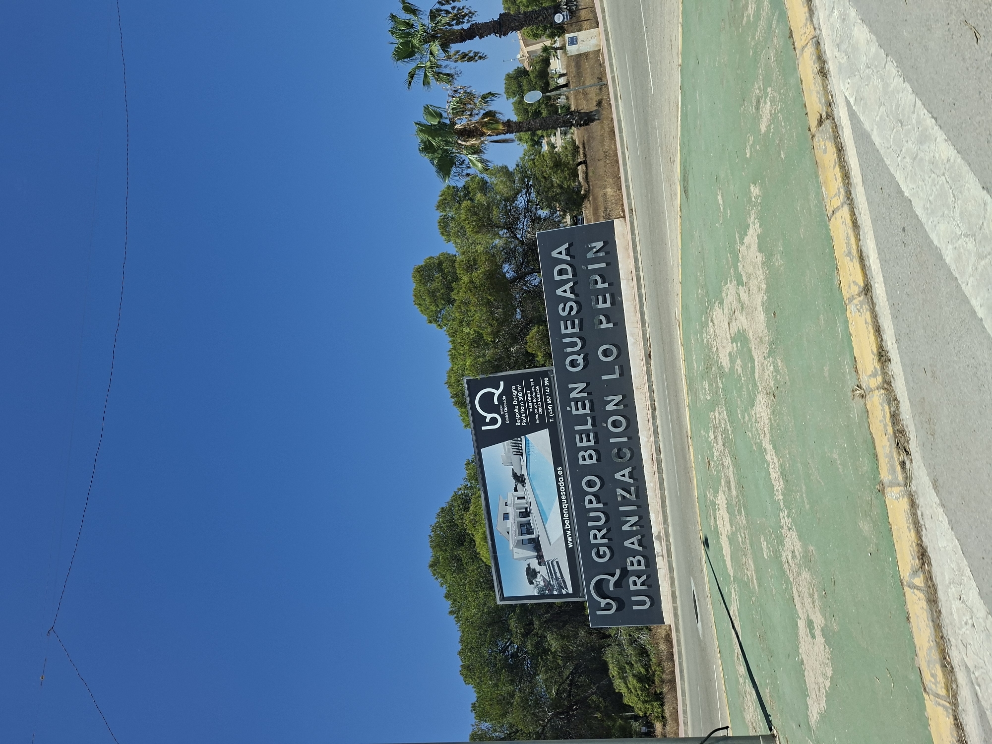 The image shows a roadside view with a clear blue sky above. In the background, a billboard features a modern house with a swimming pool, accompanied by text that reads "bQ GRUPO Belén Quesada", "Bespoke Designs Plots from 300 m²", "MAIN OFFICE Avenida de las Naciones · 18 B CIUDAD QUESADA", and "T. (+34) 657 147 390". Below this billboard, a large dark grey sign displays the text "bQ GRUPO BELÉN QUESADA URBANIZACIÓN LO PEPÍN" in raised silver lettering. The foreground shows a paved area with a green surface and a yellow curb, bordering a road. Several palm trees and lush green trees are visible behind the signs, with some residential buildings in the far distance. The location is identified as Ciudad Quesada in Rojales, Spain. No people are visible in the image.