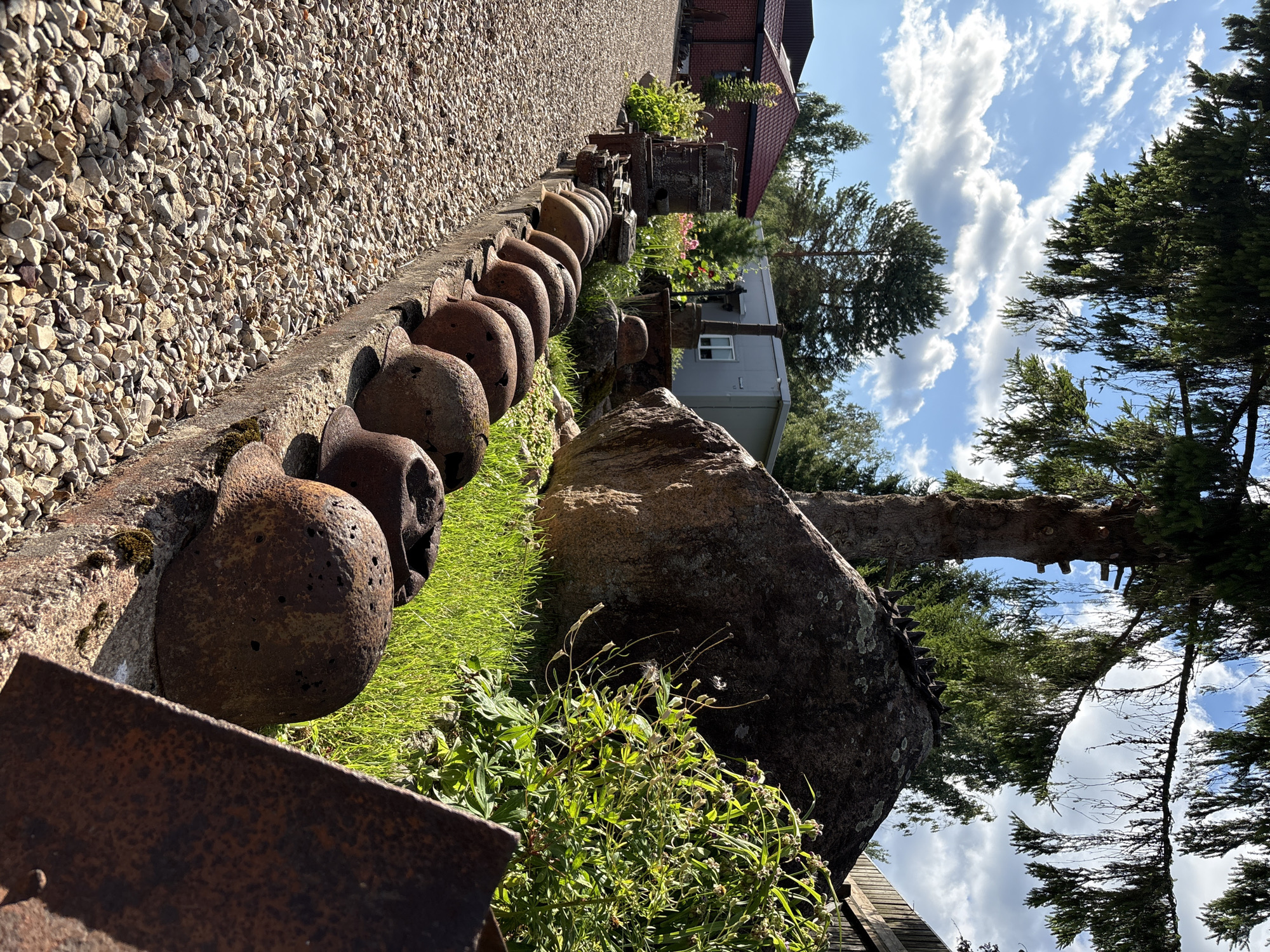 The image depicts a row of rusted metal helmets arranged along a concrete ledge. The helmets are positioned in a shallow trench, and they appear to be weathered and damaged, with visible signs of corrosion and holes. Behind the helmets, a large boulder sits on a grassy slope, partially shaded by trees. In the background, a brick building with a red roof, a modular structure, and more trees are visible. The overall scene suggests an outdoor display or memorial, possibly related to military history. The location is identified as Zhalybnya, Russia.
