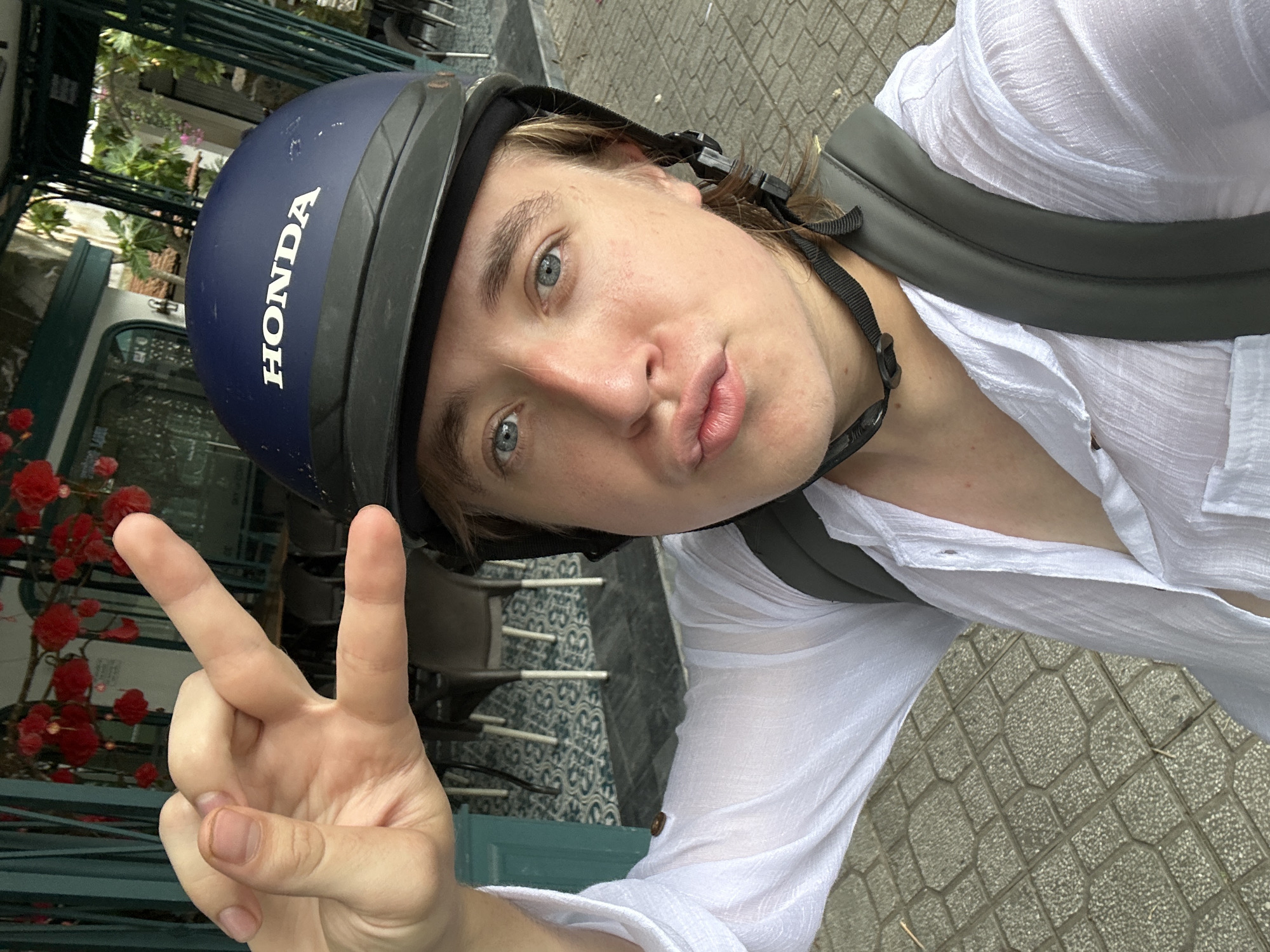 A young person with blonde hair and blue eyes, wearing a white open-collared shirt and a dark blue Honda-branded helmet, is taking a selfie outdoors. They are puckering their lips and holding up their right hand in a peace sign, with their thumb and index finger extended. The person is wearing a grey backpack. The background shows a tiled sidewalk and a building with green awnings and outdoor seating. The scene appears to be in an urban environment in Thanh Hưng, Vietnam, possibly near a cafe or restaurant, given the presence of tables and chairs. Red lanterns or decorations are visible hanging from a structure in the upper left background.