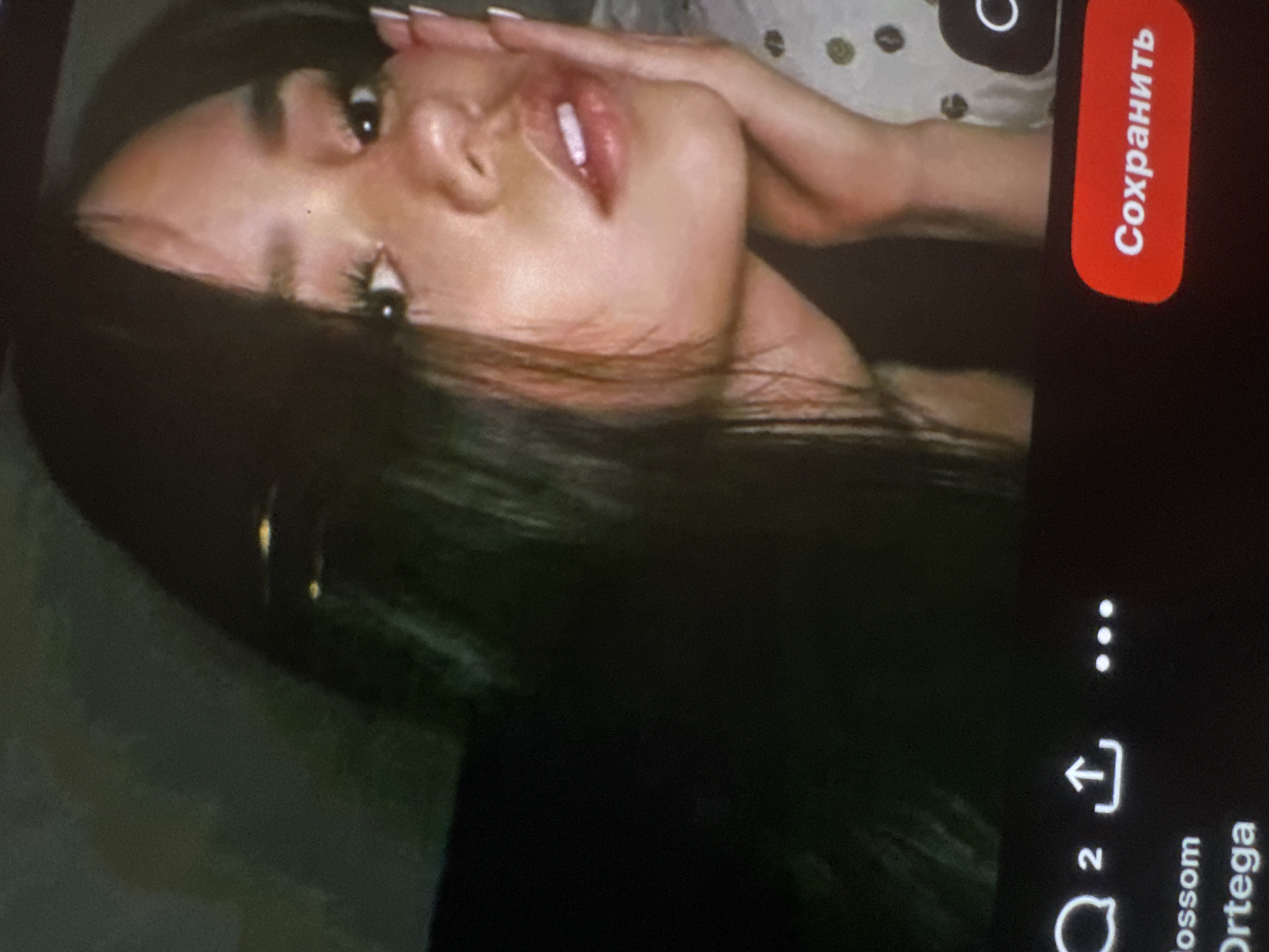 The image is a photograph of a digital screen, displaying a portrait of a young woman taken from a diagonal angle. The woman, with long, dark hair, is shown from the chest up, her head tilted. She is looking upwards and to the side, with a calm to slightly alluring expression. Her lips are slightly parted, revealing a hint of teeth and appearing glossy. She has prominent, dark eyelashes, likely enhanced. One hand, with light-colored fingernails, is visible near her face. Behind her head, a light-colored, patterned fabric with dark circular or oval shapes is visible.
The digital screen around the portrait displays elements of a user interface. On the upper right, a bright red rectangular button contains white text that reads "Сохранить," which is Russian for "Save." Below this button, against a dark background, are several social media-like icons: three vertical dots, an upload or share icon (an arrow pointing up from a box), and a speech bubble icon next to the number "2." Further down, partially visible text reads "lossom Ortega," likely part of a name or username. The image quality suggests it is a photograph taken of a screen rather than a direct digital capture, showing some screen texture and potential glare. The lighting on the woman's face is soft, and the background within her portrait appears slightly out of focus.