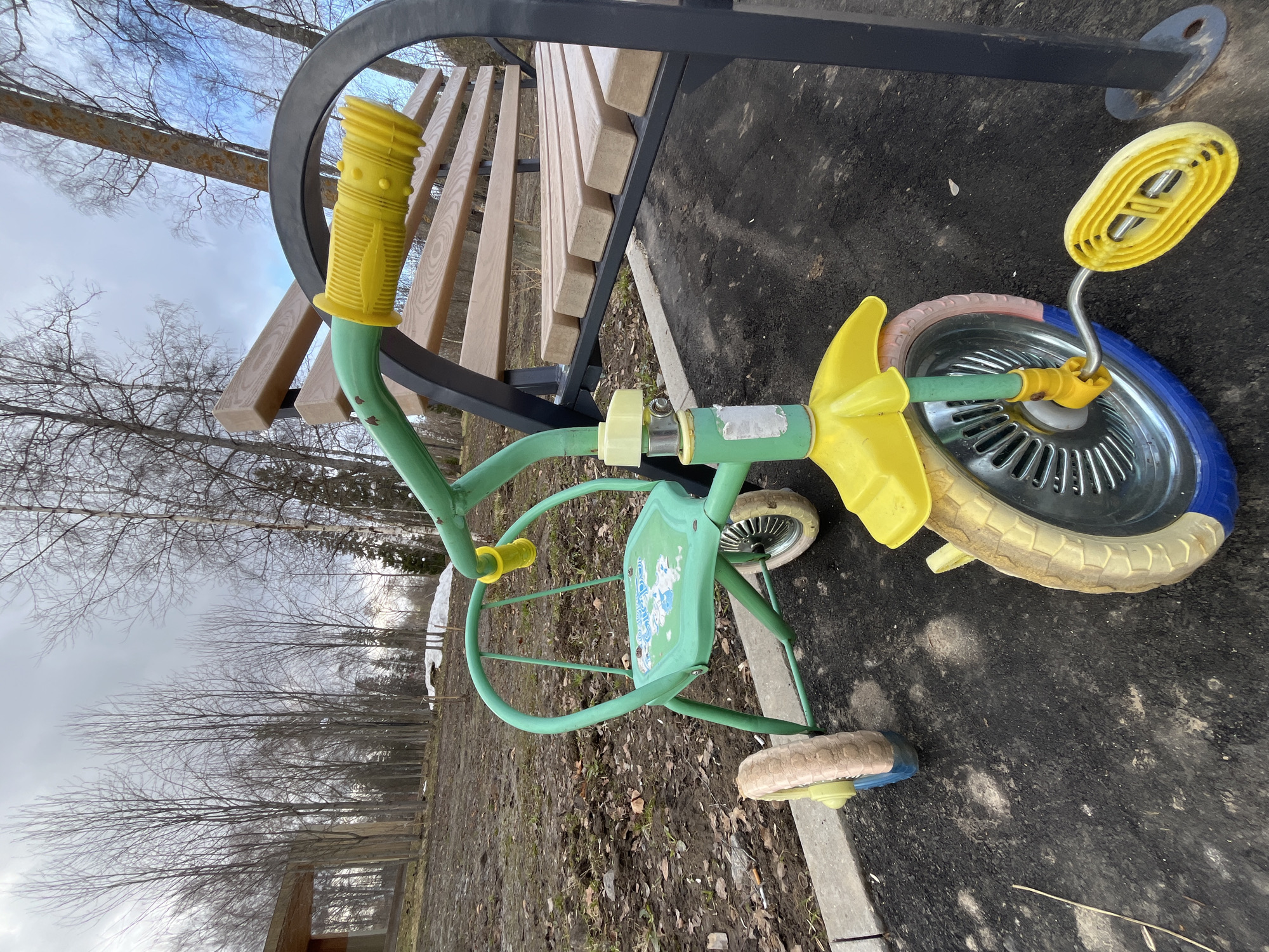 A green and yellow tricycle is situated next to a park bench in an outdoor environment. The tricycle's front wheel features a mix of colors: tan, pink, and blue on its tire. The ground around the tricycle is dark and appears to be a combination of soil, mulch, and pavement. A park bench with light-colored wooden slats and a black metal frame is partially visible behind the tricycle. Bare trees populate the background, suggesting a season such as late autumn or early spring. The sky is overcast. The location is likely in Vysokovsk, Russia.