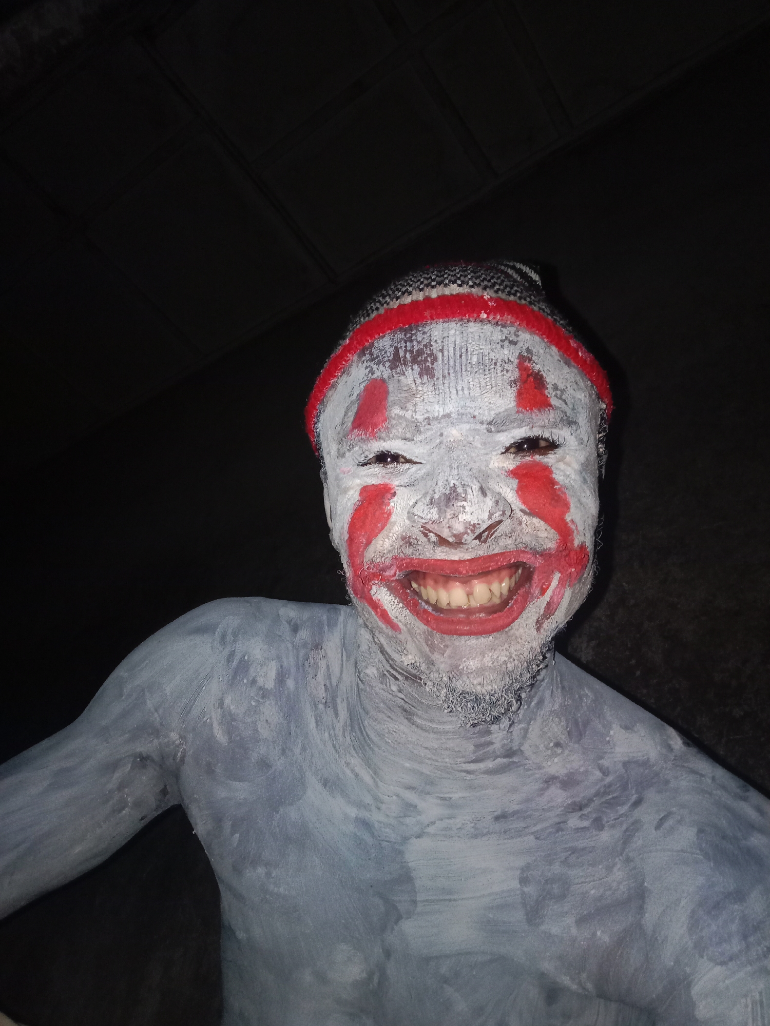 A person is shown from the chest up, with the face painted white and decorated with red markings around the eyes and mouth. The person is smiling widely, revealing teeth. A red and white knitted hat is visible on the head. The body is covered in light gray paint. The background is dark, obscuring details about the environment. The location and city are unknown.