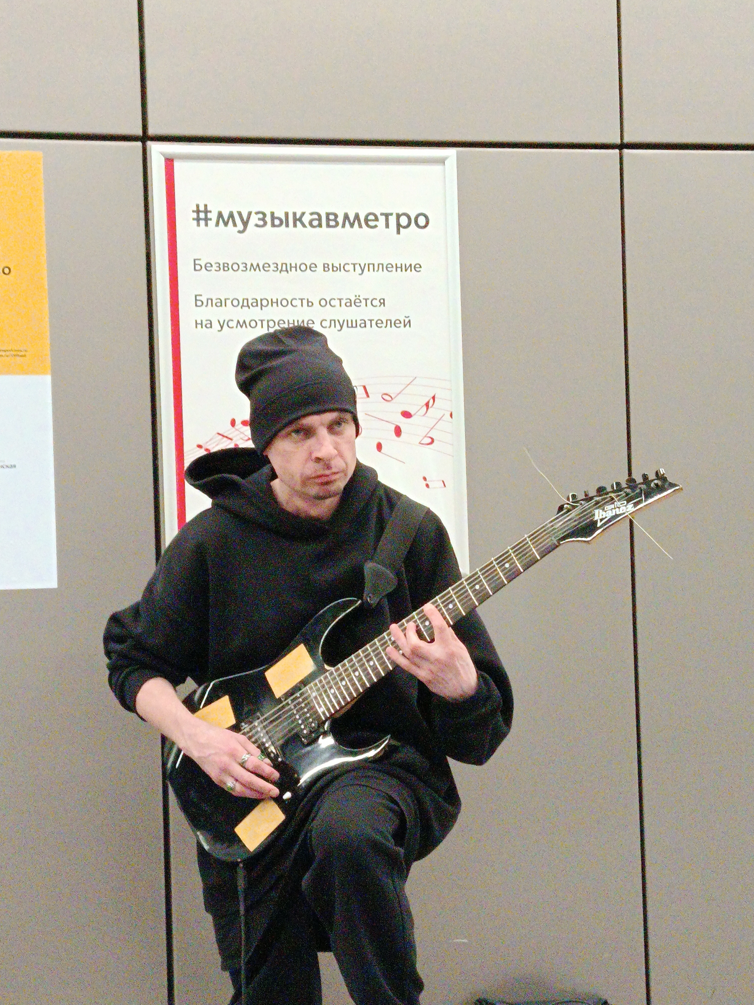 A man in a black hoodie and beanie is playing an Ibanez electric guitar in what appears to be a metro station in Moscow, Russia. The man is positioned in front of a poster that reads "#музыкавметро" (music in the metro) in Russian. The text below translates to "Free performance. Gratitude is at the discretion of the listeners." The background is a simple grey tiled wall.