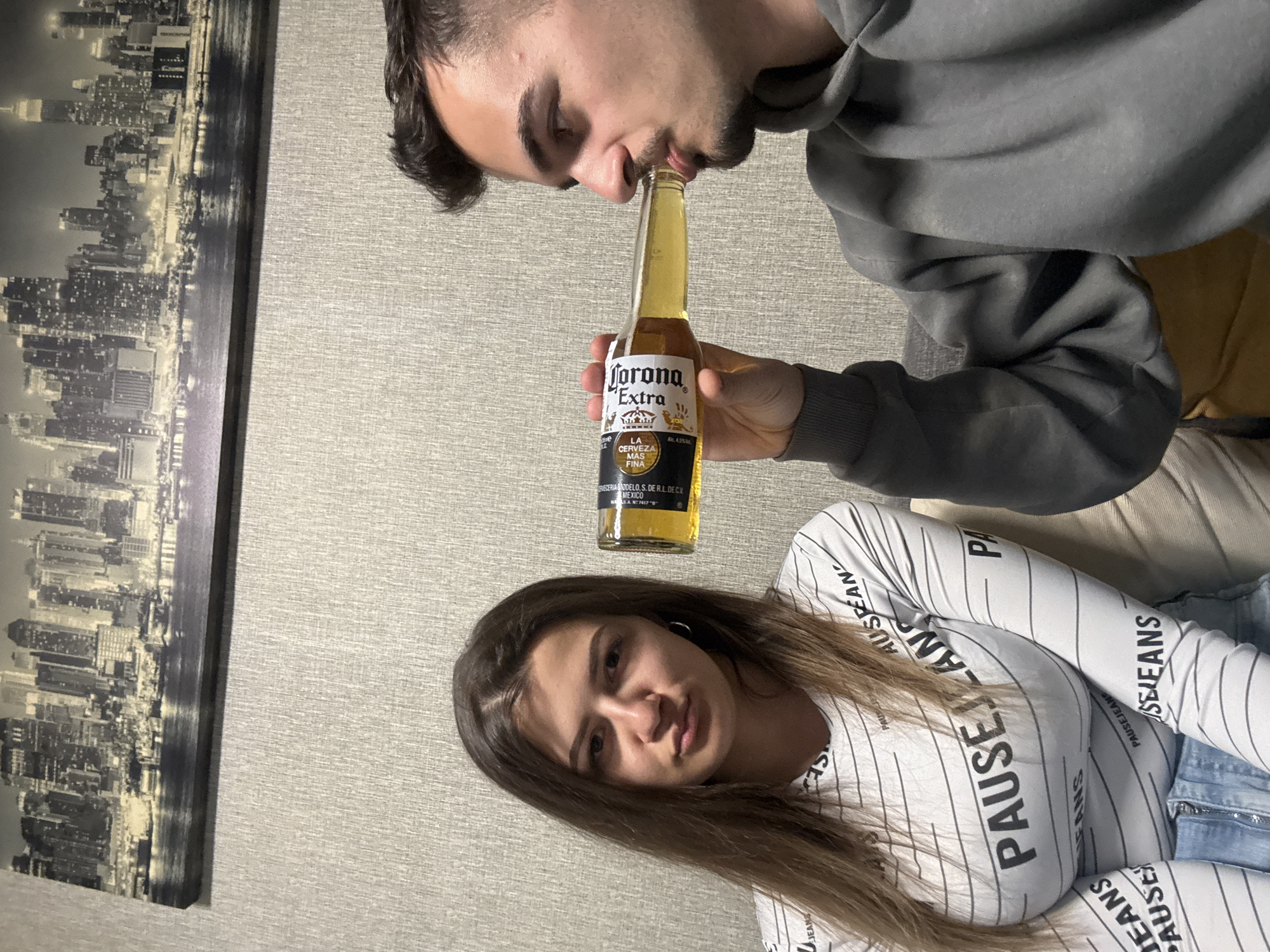 Two people are in the image. The man has brown hair and a beard, and is wearing a grey hoodie. He is drinking a Corona Extra beer. The woman has long brown hair, and is wearing a white long-sleeve shirt with the word "PAUSE" written on it, and blue jeans. There is a black and white photo of a city skyline on the wall. The location is unknown and may not be in Sofia, Bulgaria.