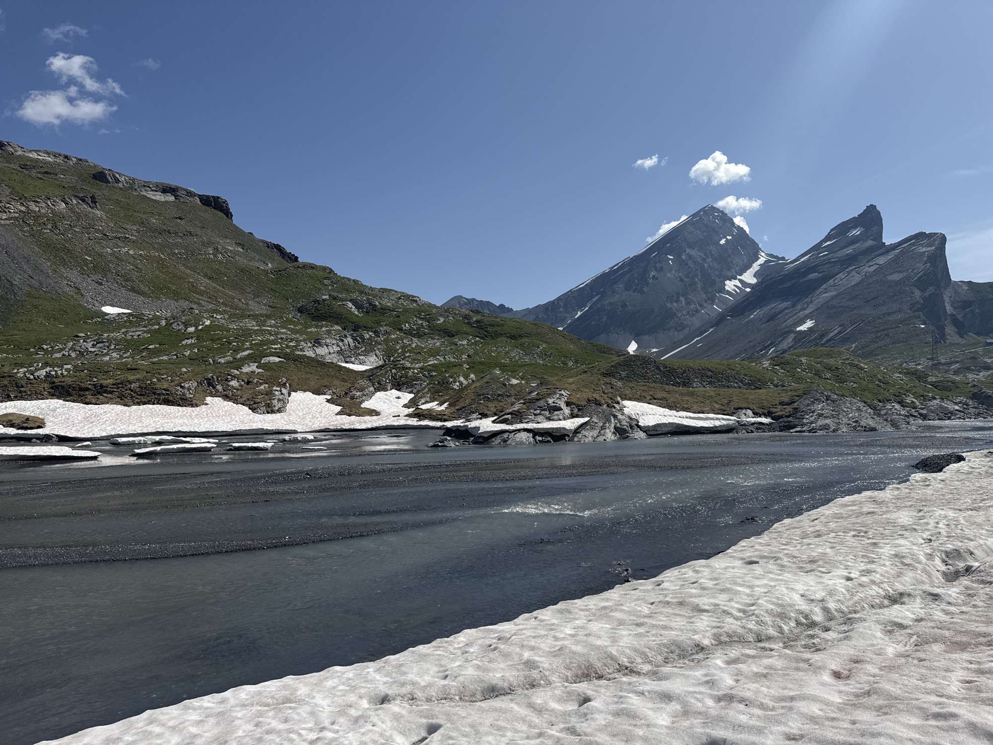 This is a landscape photograph taken in Leukerbad, Switzerland, depicting a mountainous scene with a river and snow. The sky is clear and blue, with a few wispy clouds. In the foreground, a wide, shallow river flows from left to right across the frame. The riverbed is composed of dark pebbles and sand. Snow is visible along the right bank of the river and in patches on the left bank, interspersed with rocky terrain and sparse green vegetation.
The middle ground features a rocky, green-covered mountainside rising steeply on the left, leading to a rugged ridge line. On the right, majestic, sharp-peaked mountains dominate the skyline. These peaks are dark gray rock with streaks of snow clinging to their upper slopes and crevices.
The overall environment suggests a high-altitude location during a season where snow is melting and rivers are active, likely late spring or early summer in the Swiss Alps. There are no people or animals visible in the image. The scene is natural and undeveloped.