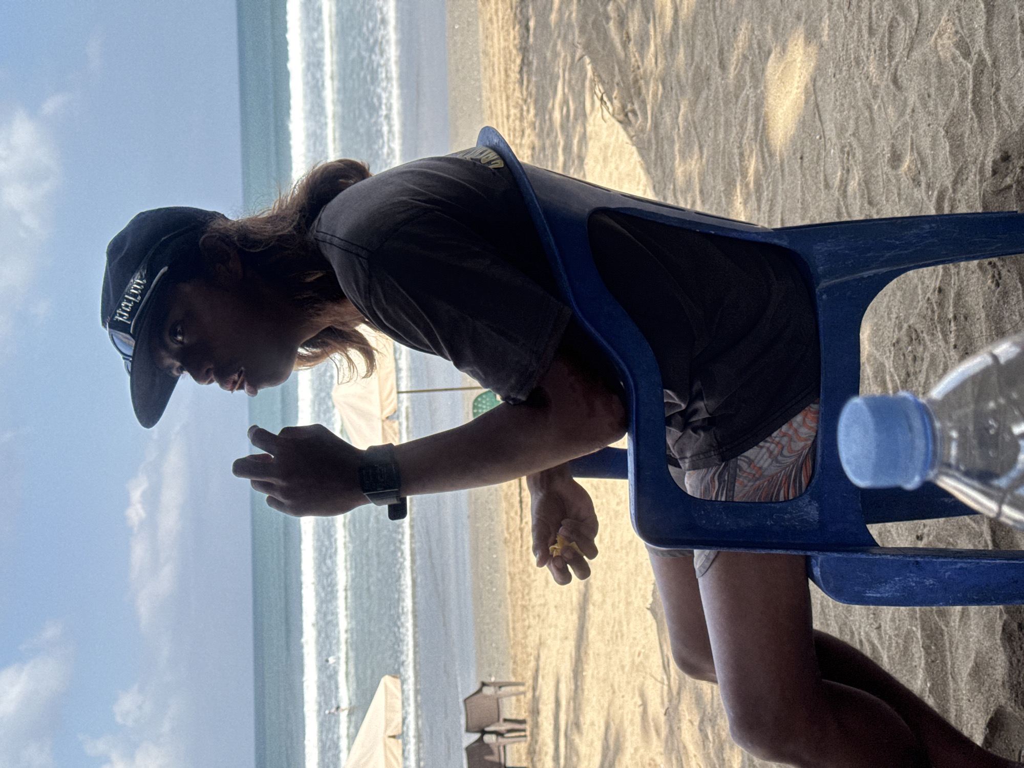 A person sits on a blue plastic chair on a sandy beach, facing the ocean. The individual is wearing a dark t-shirt, patterned shorts, and a black baseball cap. Their right hand is raised towards their mouth, and they appear to be holding a small yellow object. In the background, the ocean waves are visible, with a few beach umbrellas and chairs set up further down the shore. The sky is blue with scattered white clouds. A partially visible plastic water bottle with a blue cap rests on the sand in the foreground. The scene is likely Kuta, Indonesia, given the characteristic beach and surf.