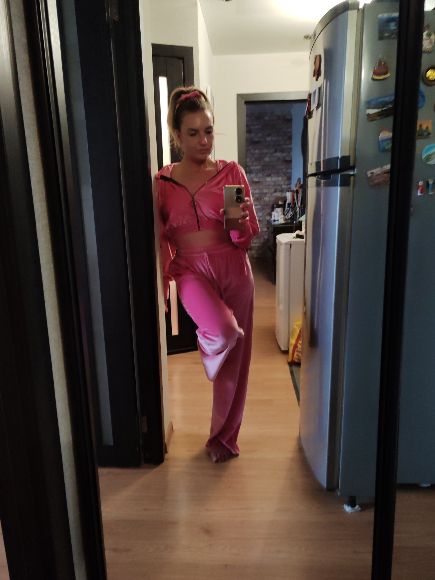 A woman is reflected in a mirror in what appears to be a hallway/kitchen area of a home. She is fair-skinned, with blonde hair pulled back, wearing a pink velour tracksuit and holding a smartphone up as if taking a selfie. The interior features a wooden floor, dark trim around doorways, a refrigerator with magnets on it, and a glimpse into another room with a brick wall. The scene is indoors. No information indicates the location is Petropavlovsk-Kamchatsky, Russia.