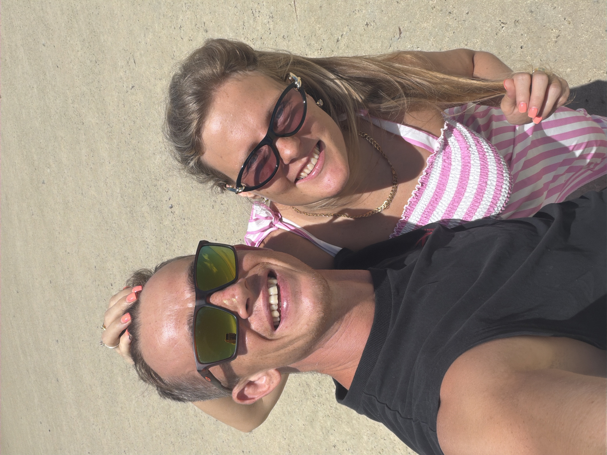 A medium shot captures a man and a woman in a selfie-style photograph against a sandy backdrop. The man, positioned on the left, has his arm raised and his hand on his head, revealing a ring on his finger. He is wearing a black t-shirt and sunglasses with orange-tinted lenses. To his right, the woman, partially obscured, is wearing a pink and white striped dress and sunglasses. A gold chain necklace is visible around her neck, and her fingers are touching the dress fabric. The environment appears to be a beach or a sandy area, likely in ตำบลเกาะพะงัน, Thailand, given the context. The lighting suggests it is daytime, with bright sunlight casting shadows.