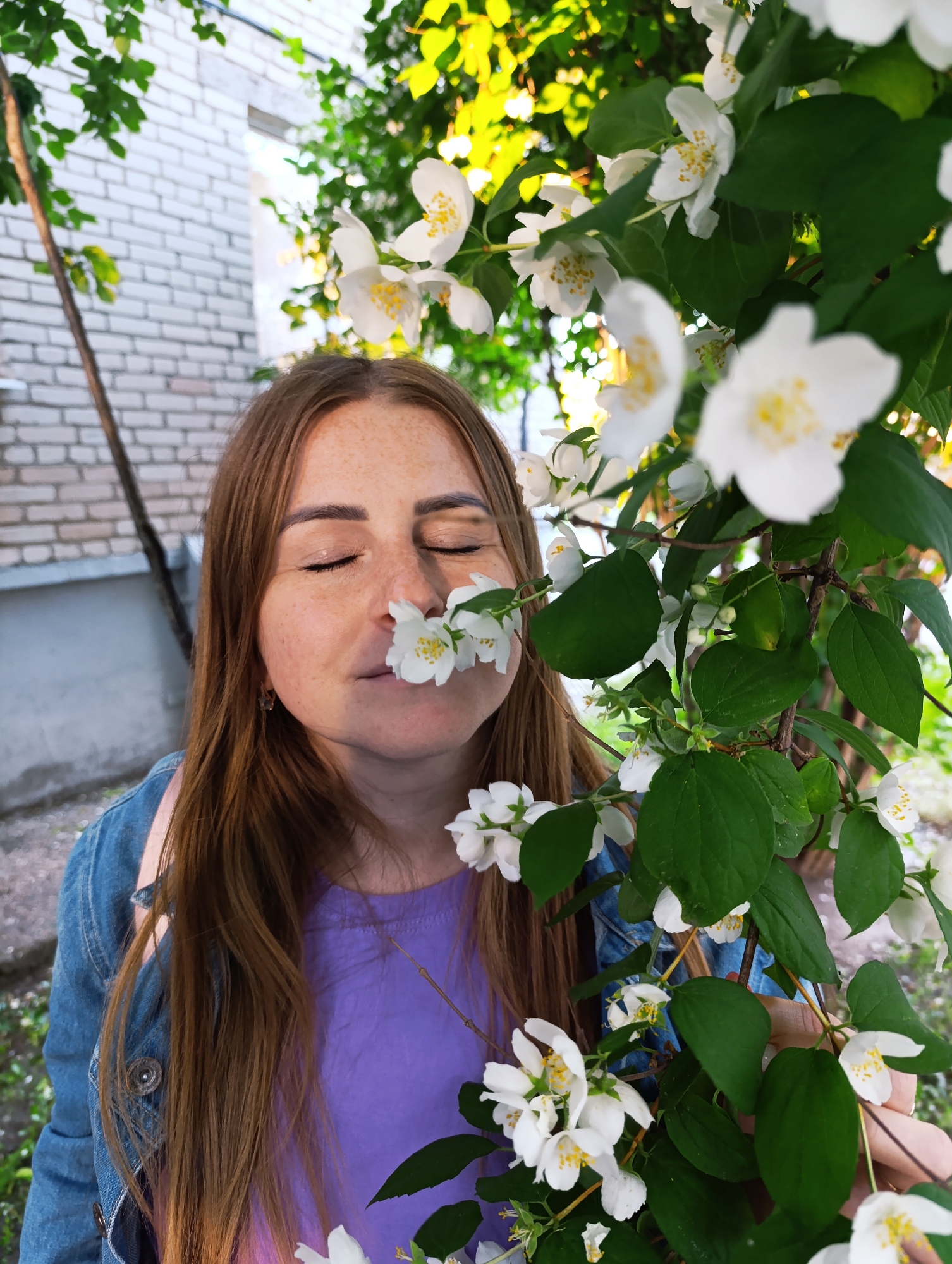 A woman with long brown hair and freckles is seen from the chest up, in a medium shot, outdoors. She is wearing a denim jacket over a purple t-shirt. Her eyes are closed, and she is holding a sprig of white flowers, which she is smelling by bringing them close to her nose. The flowers are delicate with yellow stamens in the center. Behind her, a light-colored brick wall is visible, along with green foliage from trees and bushes. Sunlight filters through the leaves, creating a bright, possibly late afternoon or early morning, outdoor scene. The overall mood is peaceful and serene. The location is identified as Chernogolovka, Russia.