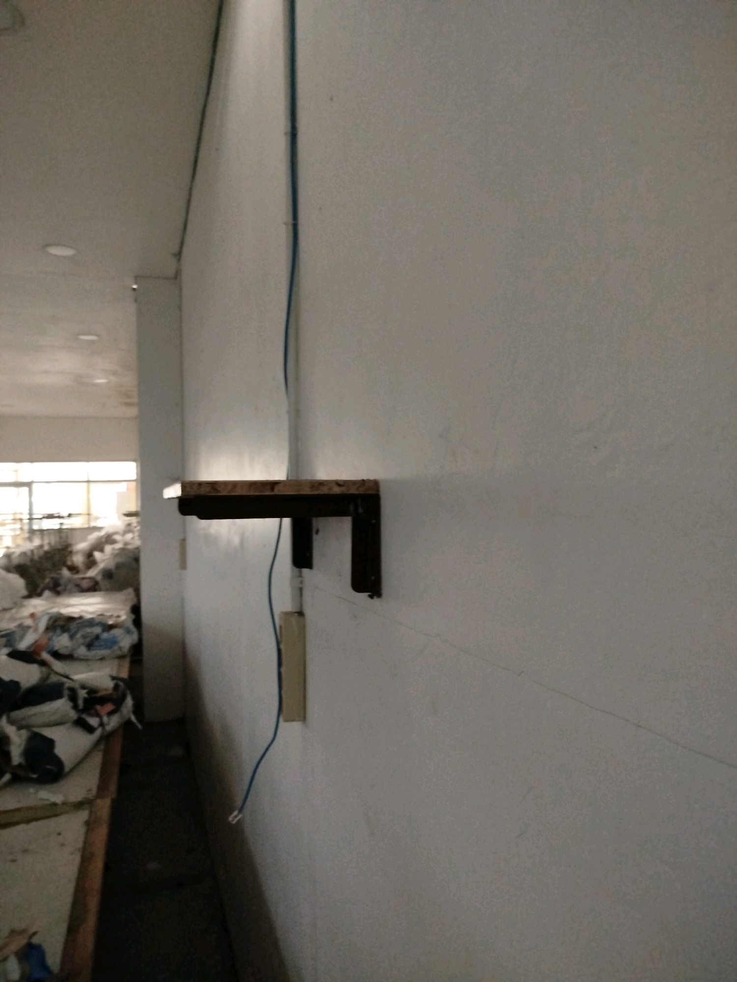 The image depicts an indoor scene, possibly a workshop or storage area, with a focus on a wall-mounted shelf. The shelf is made of wood with a dark, possibly metal, bracket. A thin blue wire runs vertically along the wall, passing behind the shelf and extending downwards to an electrical outlet box. To the left of the wall, a hallway or aisle is visible, lined with stacks of what appear to be fabric or textile materials, some of which are bundled and covered. The flooring in this aisle is dark and appears somewhat worn. The walls are painted a light, neutral color, showing some texture and subtle lines. Natural light filters in from a window or opening at the far end of the aisle, illuminating the stacked materials. The overall impression is one of an industrial or utilitarian space. The image does not contain enough information to definitively place it in Surakarta, Indonesia.