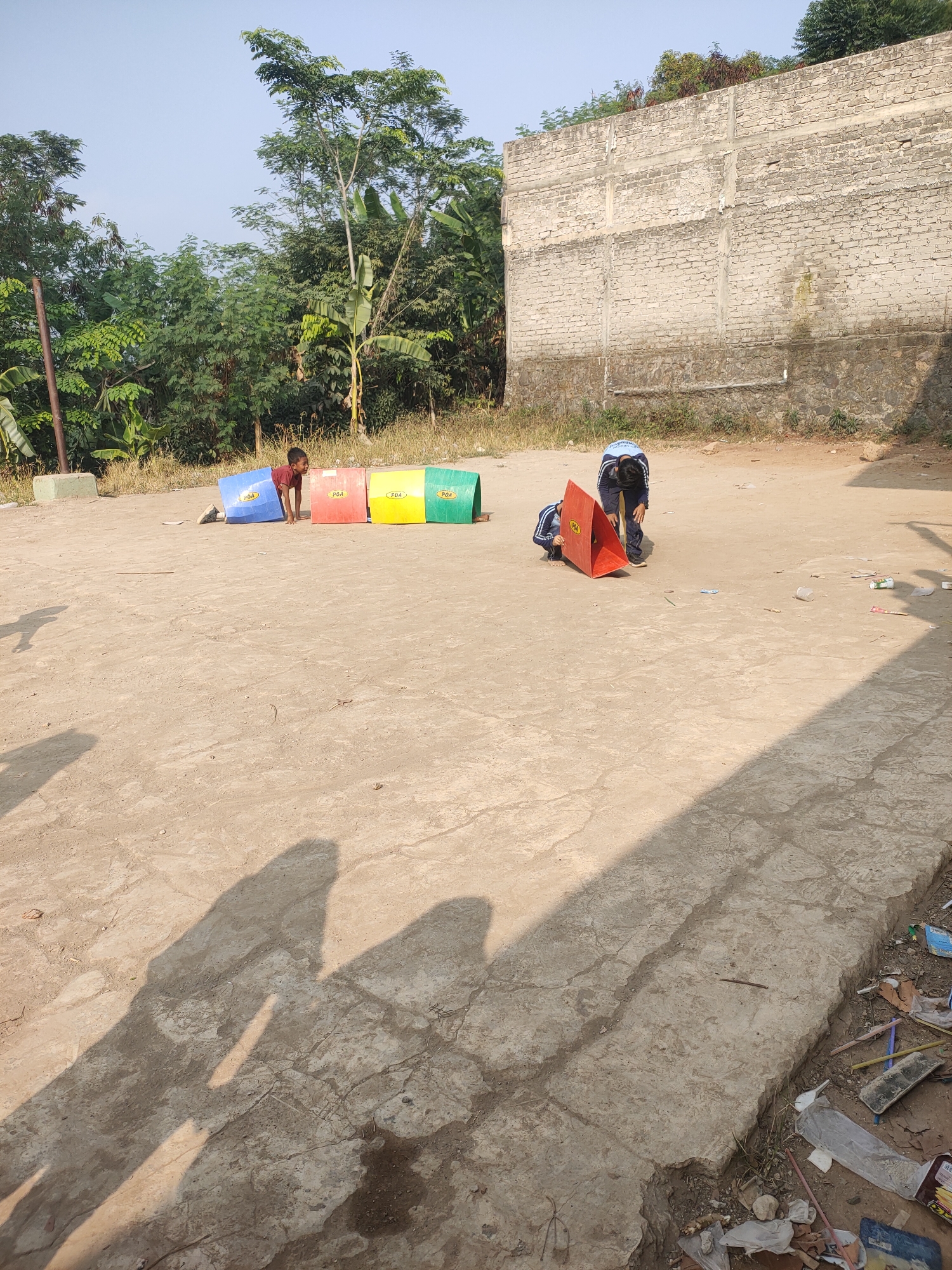 In Subang, Indonesia, on a dirt and concrete lot, two young boys are engaged in an outdoor activity. One boy, wearing a red shirt, crawls inside a blue cylindrical obstacle. To his right, a red, yellow, and green obstacle stands in a line. Further to the right, a second boy, dressed in a blue and white uniform, bends over a red cone-shaped object. The scene is bathed in sunlight, casting distinct shadows on the ground. In the background, a weathered brick wall and lush greenery suggest a semi-urban or rural environment. Scattered debris, including plastic bottles and wrappers, are visible on the ground.