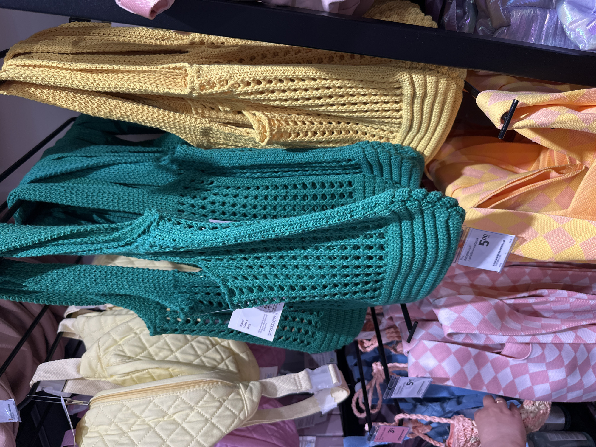 A retail display features an assortment of handbags and bags on a black metal rack. In the foreground, two large, crocheted tote bags are prominently displayed. The bag on the left is a vibrant teal color, and the bag on the right is a bright yellow. Both bags have an open weave pattern with larger holes and a more tightly woven band at the bottom. Behind these, a cream-colored, quilted fanny pack is visible, along with a portion of a pink bag. Further down, there are several patterned tote bags in pink and yellow, as well as a small pink woven bag. Price tags are attached to some of the items. The background is blurred, showing more merchandise and shelving. The lighting suggests an indoor retail environment, possibly a department store or boutique.
