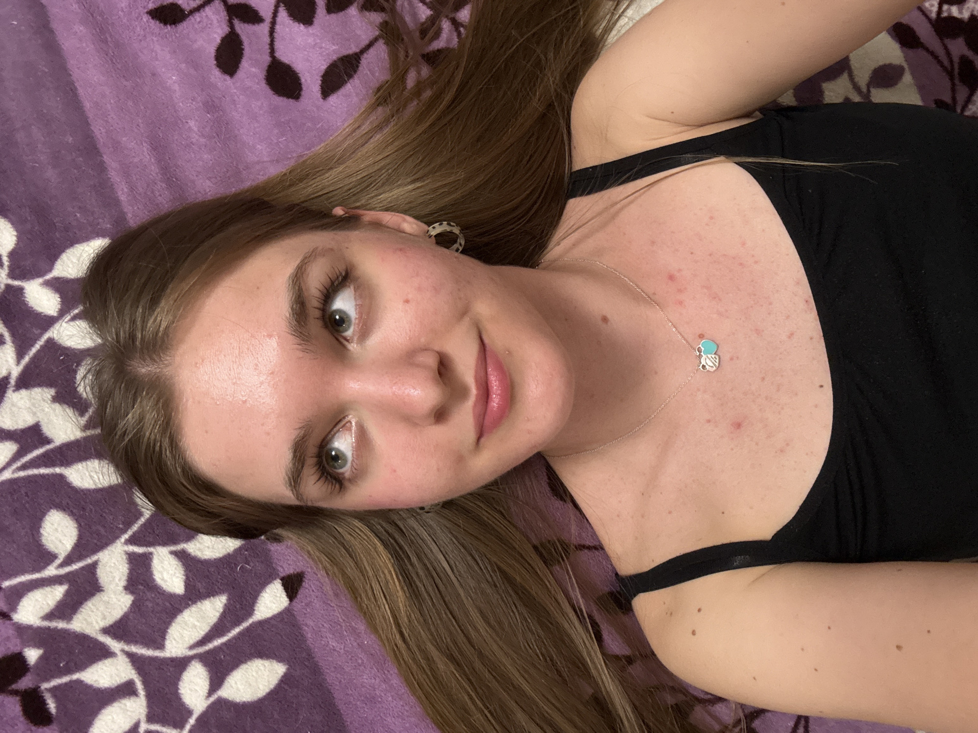 A young woman is lying on a patterned purple blanket or bedspread. She is wearing a black tank top and a silver necklace with a blue pendant. Her hair is long and brown, and she is wearing patterned earrings. The scene does not provide any indicators of the location in Perm, Russia.