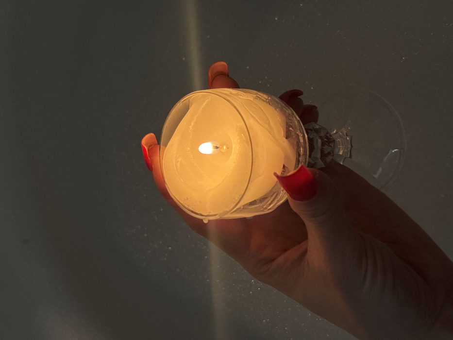A human hand, likely female, with long red-painted fingernails, holds a clear glass stemmed goblet containing a lit white candle. The candle's flame is bright, casting a warm yellow-orange light that illuminates the hand, the wax, and the inner surface of the goblet, which shows solidified, irregular wax patterns. The goblet has a faceted base and stem. The background is dark and indistinct, revealing faint, blurred highlights consistent with the surface of water with small bubbles or foam, suggesting a bath environment. The scene is located in Чистенское сельское поселение, Ukraine.