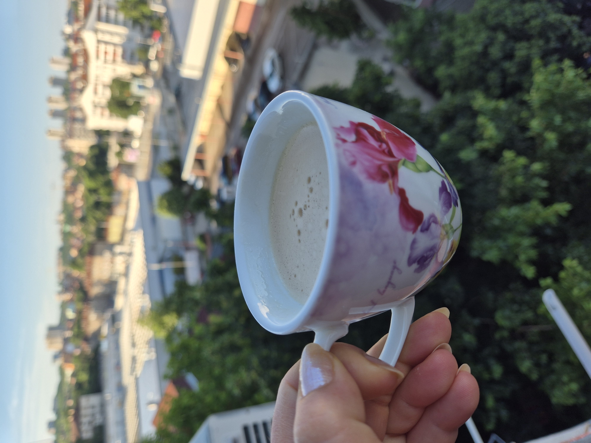 A hand is holding a decorative cup filled with a frothy beverage against a blurred cityscape and lush green trees. The cup features floral designs in shades of pink and purple. The background shows buildings of various sizes and colors under a clear sky, suggesting an urban environment. The focus is on the cup and hand, with the city serving as a backdrop.