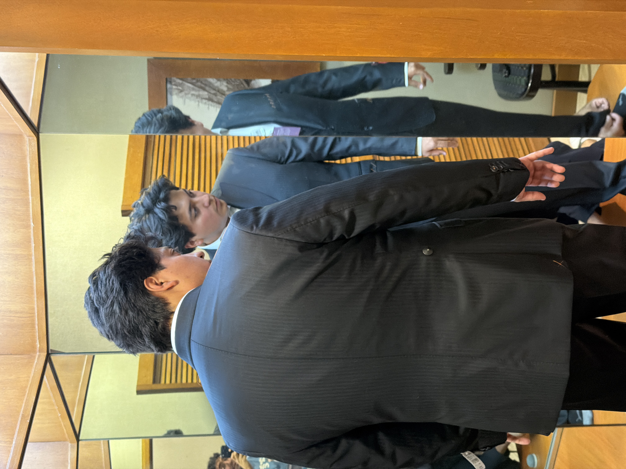 A young man wearing a black suit stands in front of a multi-paneled mirror. The reflection shows the man fully, revealing that he is also wearing a light-colored shirt with a possible purple tie. The mirror appears to be part of a wooden structure, likely a cabinet or display. The location is the city of Puebla, Mexico.