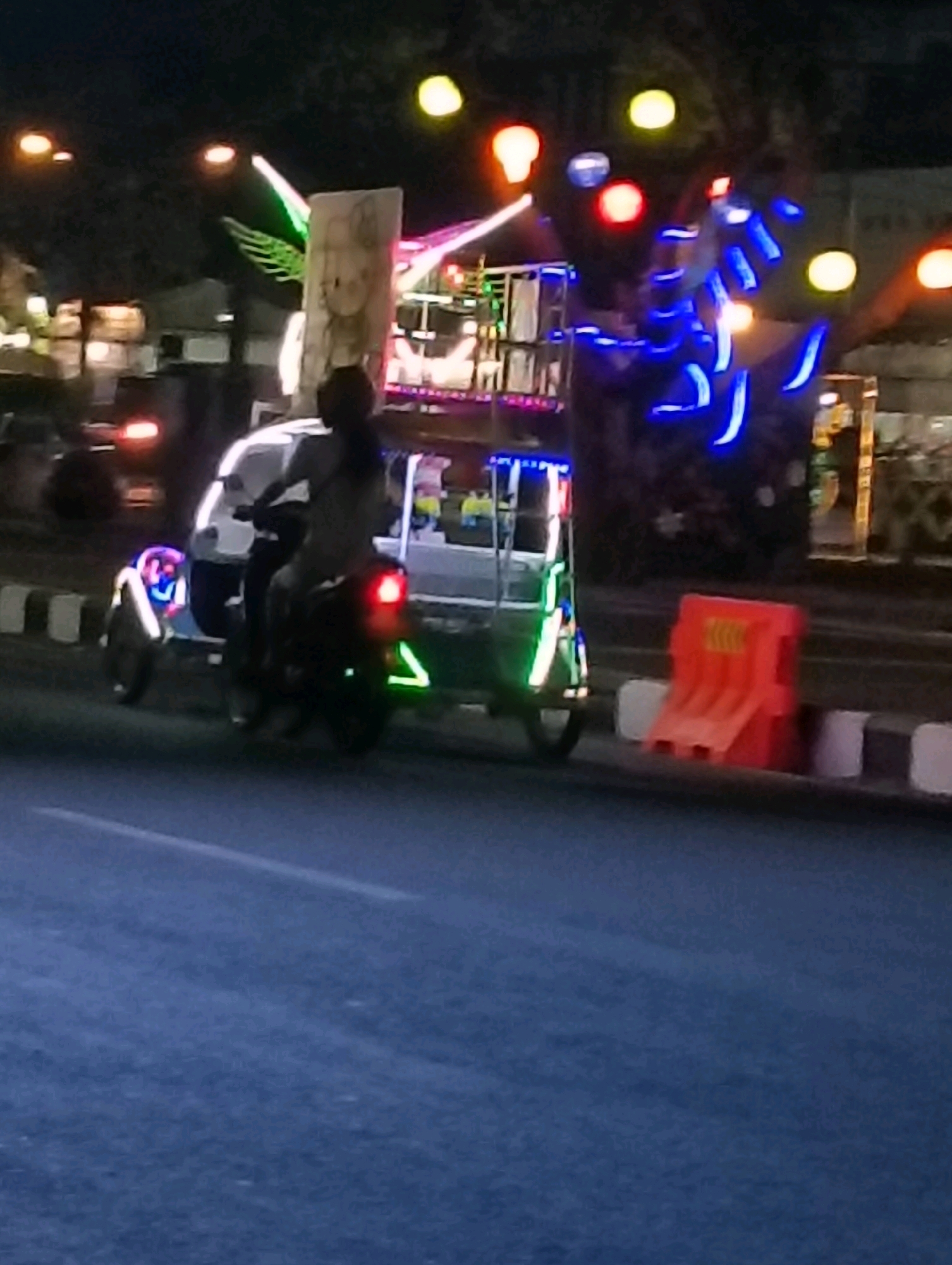 The image depicts a nighttime scene on a road in Purworejo, Indonesia, featuring a brightly lit three-wheeled vehicle, likely a modified becak motor, adorned with an array of colorful LED lights, including blue, green, and red, and possibly some reflective tape on its sides. A person, presumably the driver, is seated on the vehicle, facing forward. The vehicle carries a structure on its back, possibly for entertainment or transport, with more lights and a depiction of a cartoonish character on a sign. In the background, there are streetlights and further illuminated structures, suggesting a commercial or entertainment area. On the side of the road, an orange traffic barrier is visible. The road surface appears to be asphalt, with white and black striped road markers. The overall ambiance is festive, illuminated by artificial lights against the dark night sky.