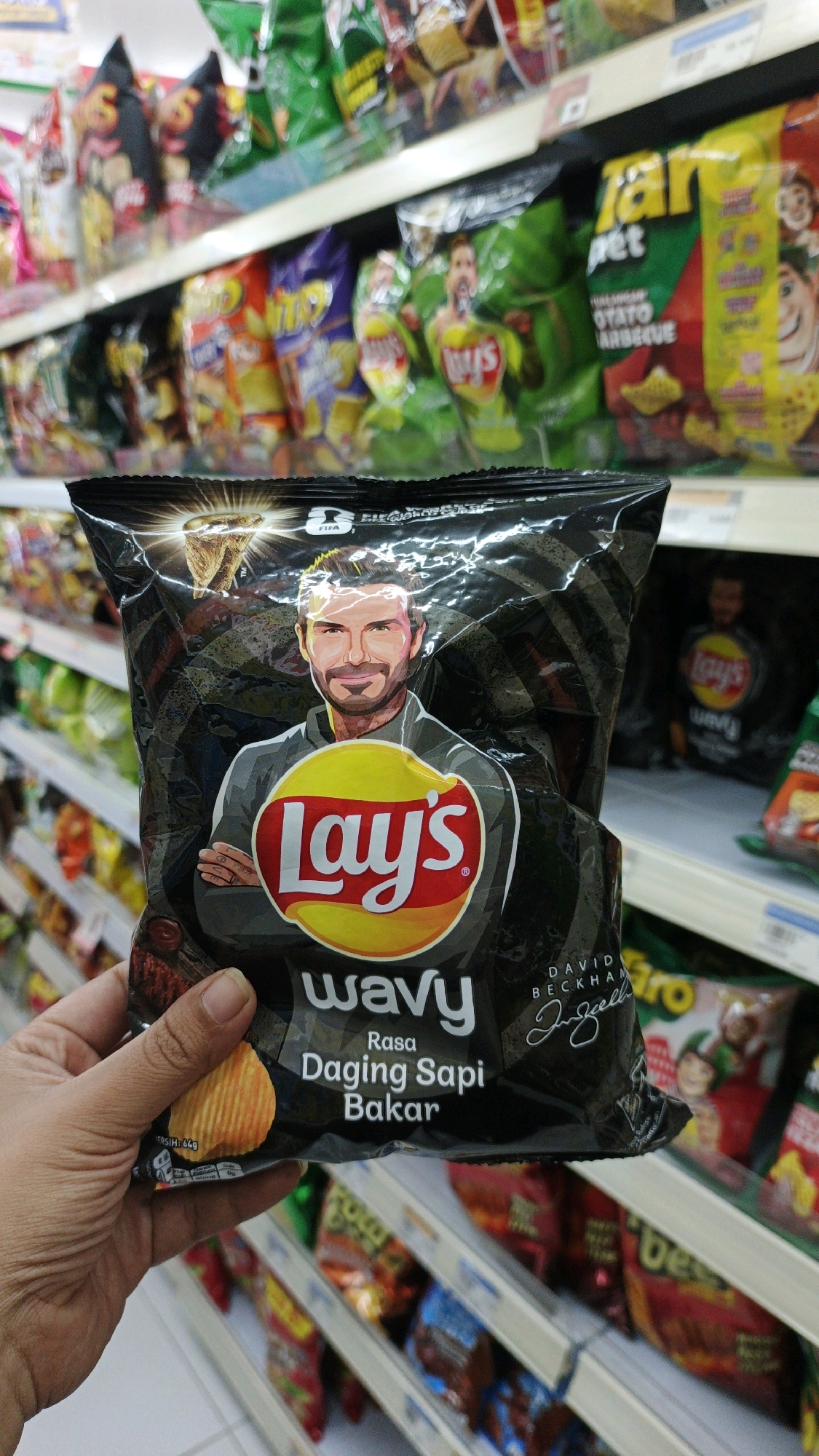 A person's hand holds a black bag of Lay's Wavy potato chips featuring an illustration of David Beckham. The chips are described as "Rasa Daging Sapi Bakar" (Barbecued Beef Flavor). The background shows shelves stocked with various snacks and chip bags in a brightly lit supermarket aisle. This scene is consistent with a retail environment, potentially a minimarket or supermarket, and such a display is common in urban and semi-urban areas of Indonesia, including the city of Parigi, where consumer goods are widely available in such establishments.