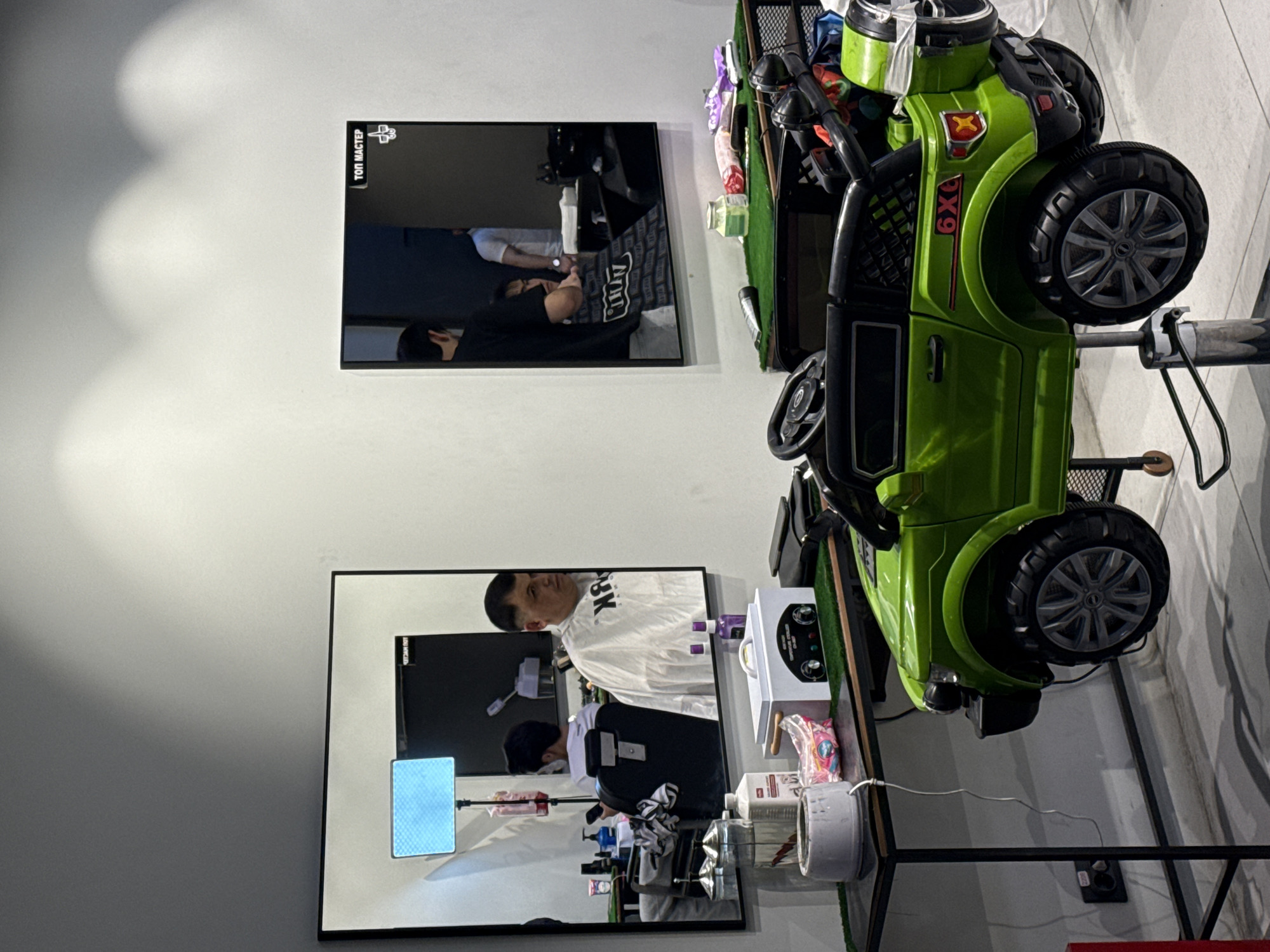A brightly lit barbershop interior is depicted, with two large mirrors reflecting the scene. In the foreground, a green toy electric ride-on car, designed to resemble an SUV with "6X6" branding, serves as a barber chair. The car has realistic-looking wheels, headlights, and a spare tire mounted on the back. It is raised on a barber chair mechanism, allowing for height adjustment.
To the left, a black metal table with a green turf-like surface holds various barber supplies, including a wax warmer, bottles of liquid, and packaged items. A person in a white cape is seated in a barber chair in the reflection of the left mirror, being attended to by a barber in a black shirt. Another person, also in a white shirt, is visible in the background reflection of the same mirror.
To the right, another mirror displays the reflection of a barber in a black t-shirt, working on a customer seated in a barber chair. The customer is draped with a cape. Above this mirror, a sign in Russian reads "ТОП МАСТЕР" (TOP MASTER). The walls are painted a light color, and the lighting is consistent with an indoor commercial establishment. The overall impression is a modern and child-friendly barbershop, likely in Moscow, Russia, based on the Russian text.