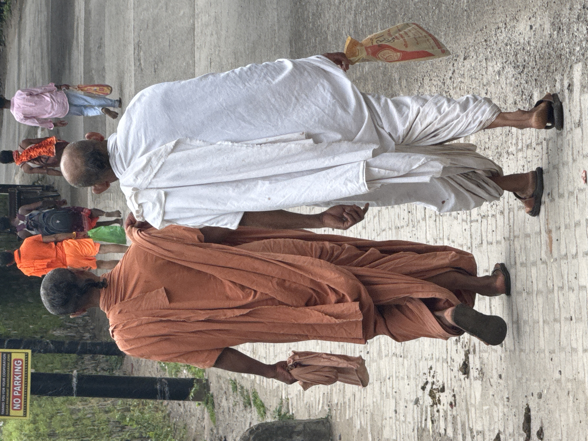 The image depicts two men from behind, walking on a paved path in Rishikesh, India. The man on the left is wearing an orange robe and has a small braid in his hair. He is carrying a folded cloth in his left hand. The man on the right is wearing a white dhoti and a white shawl draped over his shoulders. He is carrying a colorful bag in his right hand. In the background, other individuals are walking away from the camera, one wearing an orange garment and shorts, another in a pink shirt and light blue pants, and a person with a backpack. A "NO PARKING" sign is visible on the left side of the image. The environment appears to be an outdoor urban setting with some greenery on the left.