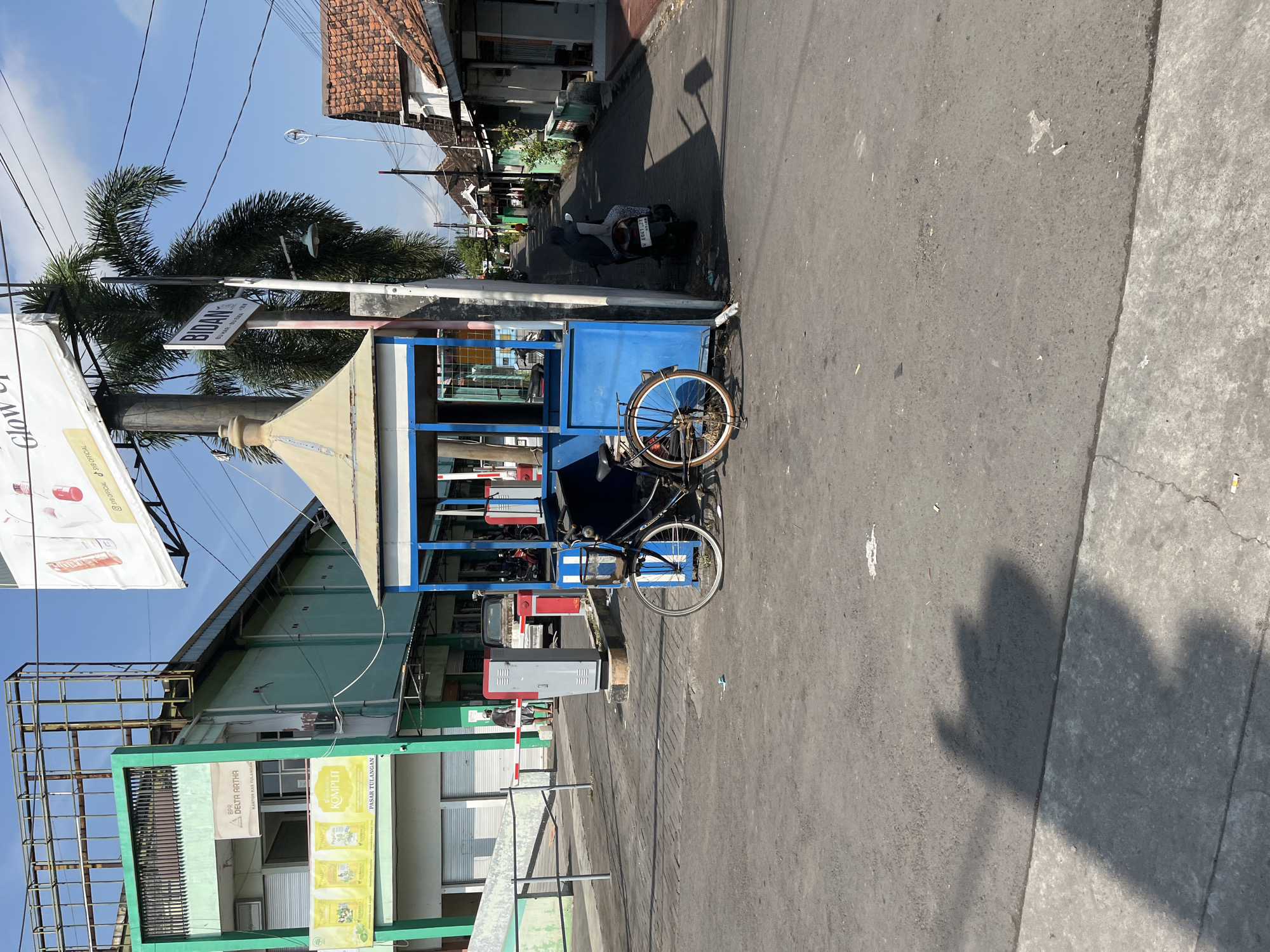 The image depicts a street scene in Tulangan, Indonesia, on a sunny day. In the foreground, a dark-colored bicycle with a brown seat and white-rimmed wheels is parked next to a small blue and white guardhouse. The guardhouse has a peaked, yellow roof and blue siding with white trim. A sign on a pole behind the guardhouse reads "BIDAN," which translates to "midwife" in Indonesian.
To the left, a light green building with a rusted metal frame structure is visible. It has signs in Indonesian, including one that says "BPR DELTA ARTHA." A silver and red automated gate system is also present on the left side of the road.
Further down the street, on the right, are buildings with red-tiled roofs, typical of residential areas. A few motorcycles are parked along the right side of the street, and one person is seated on a motorcycle, wearing a dark hooded jacket and patterned leggings. The road surface is asphalt, and there is a clear blue sky with some clouds. Shadows are cast on the ground, indicating the direction of the sun. The environment appears to be a mixed commercial and residential area within the town.