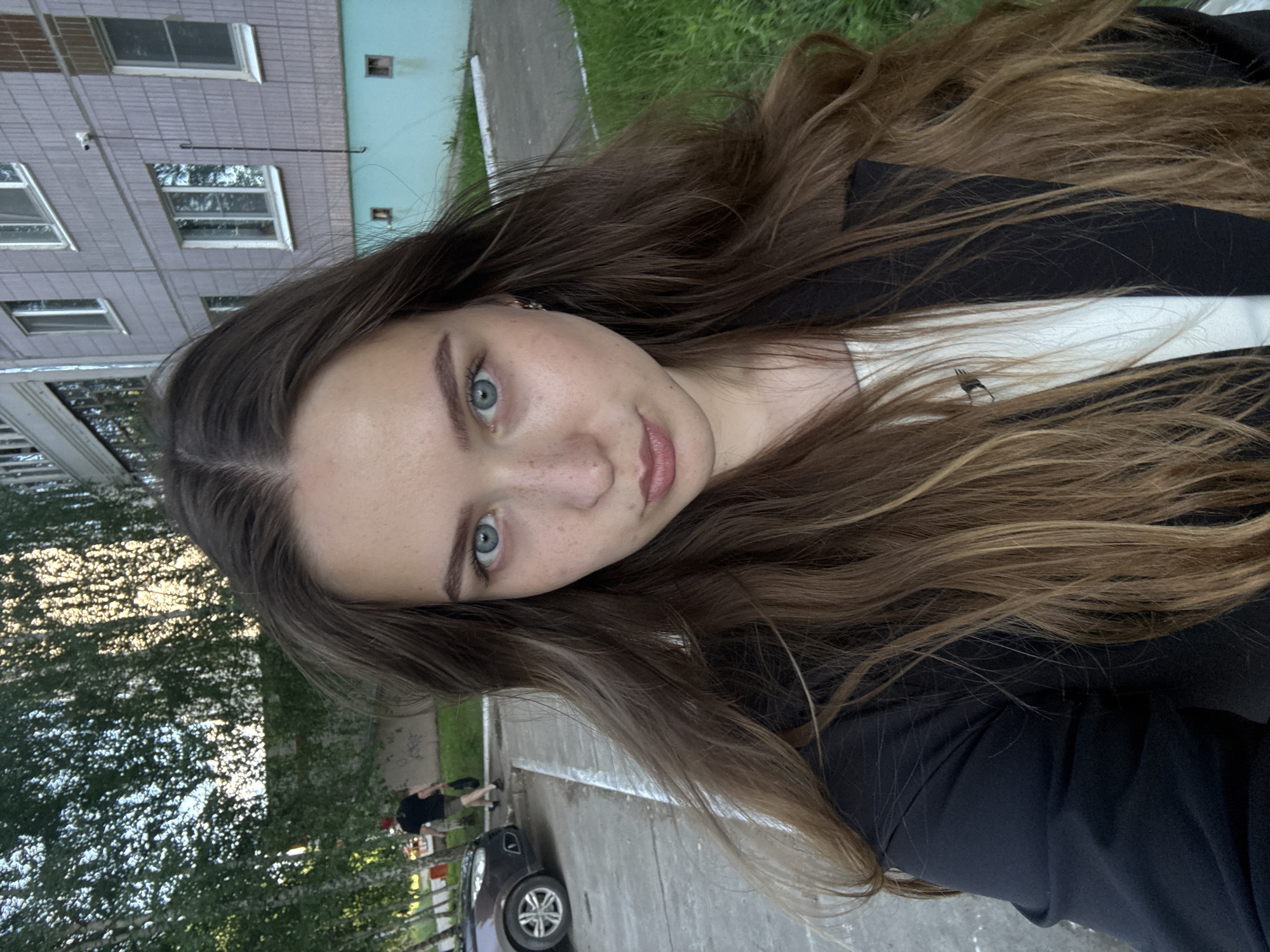 A young woman with long, light brown hair and blue eyes is pictured in a selfie taken outdoors in Chaikovsky, Russia. She is wearing a black blazer over a white top. Her facial expression is neutral, with her lips slightly pursed. Freckles are visible on her cheeks and nose.
In the background, a multi-story apartment building with gray siding and light-colored window frames is visible. A teal-colored base is present on the lower portion of the building. Green grass lines the front of the building, and a paved walkway runs along it. To the left of the woman, a dark-colored car is parked on a paved area. Further in the background, trees with green foliage line the street, and a person wearing a red hat and dark clothing is walking away from the camera. The scene appears to be an urban residential area.