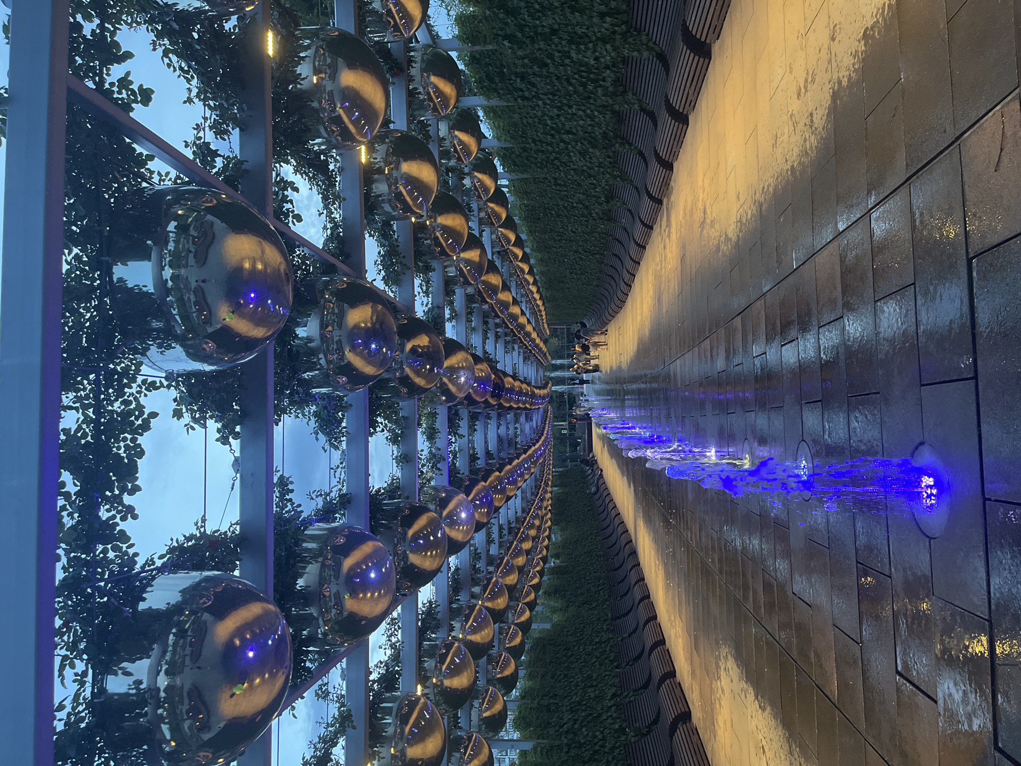 This is an outdoor scene in the city of Krasnodar, Russia. The image is taken at what appears to be an outdoor public space, featuring an illuminated fountain running along a tiled pathway, with evenly spaced blue lighting. On one side of the path, there is a wall covered in greenery, with seating. The other side features a metal structure lined with spherical metallic ornaments, reflecting the surrounding environment. There are people walking in the distance near the vanishing point of the pathway. The overall atmosphere is modern, reflecting an urban design aesthetic.