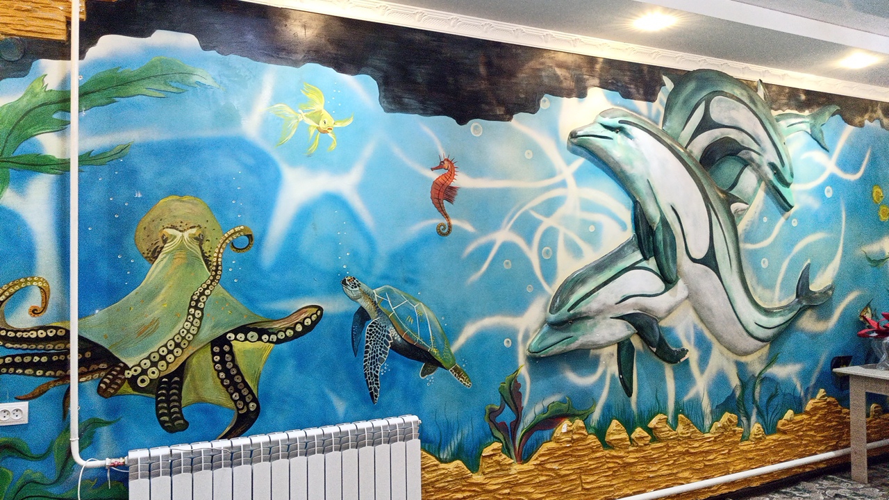 The image is a photograph of a mural on a wall in the city of Balykchy, Kyrgyzstan. The mural depicts an underwater scene with various marine life, including an octopus, a sea turtle, a seahorse, a yellow fish, and several dolphins. The mural has a textured appearance, suggesting it might be a relief mural. The background of the mural is a vibrant blue, representing water, with white swirls that suggest light filtering through the water. The lower portion of the mural features a sandy seabed with some underwater plants. To the left of the mural, a white pipe runs vertically along the wall, and a white electrical outlet is visible. Below the mural, a white radiator is installed. The ceiling above the mural has white trim and is illuminated by recessed lighting. The overall scene is a colorful and detailed artistic representation of an aquatic environment.