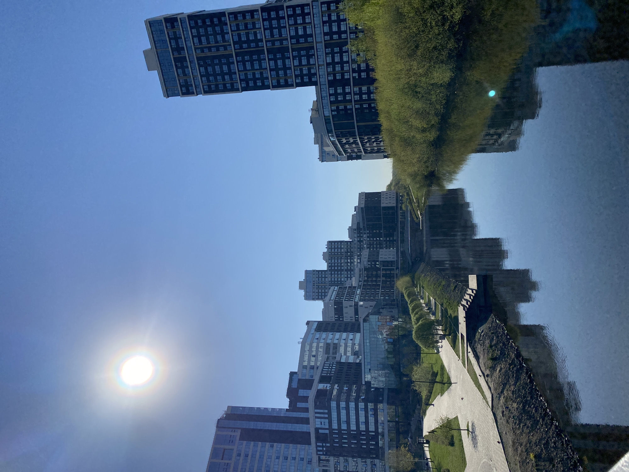 The image captures an urban landscape in Yekaterinburg, Russia. The photograph shows a row of modern high-rise buildings reflected in a body of water, likely a canal or a wide section of the Iset River, which runs through the city. The buildings exhibit a mix of architectural styles, with glass facades and varying heights. A paved walkway runs alongside the water, bordered by greenery including trees and grass. The sky is clear and blue, and the sun is positioned on the left, emitting a bright glare. The image is vertically oriented, with the actual structures appearing on the lower half of the frame and their reflections filling the upper half.