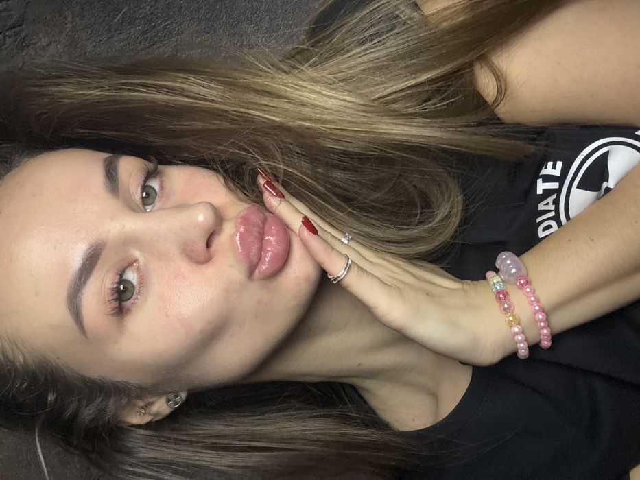 A woman with light brown hair and green eyes is the focus. Her lips are full and glossy. She wears red nail polish and two rings. Her hand rests near her face. She wears two beaded bracelets, one pink and one with multicolored beads. She is wearing a black t-shirt, part of which is visible.