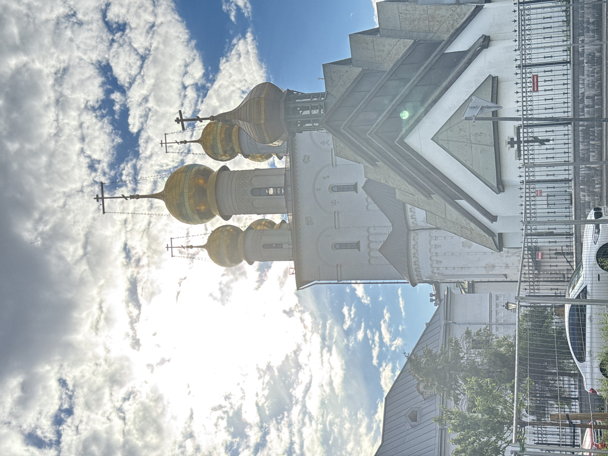The image, taken in Saint Petersburg, Russia, showcases a segment of a grand, white cathedral with distinctive golden domes, reflecting the bright sunlight. The cathedral's architecture features ornate detailing on its upper sections, with multiple onion domes topped with crosses. These domes are arranged in a cluster, suggesting a significant religious structure. The building's facade is detailed with windows and decorative elements.
In the foreground, a modern metal fence, possibly indicating a construction or maintenance area, partially obscures the view of the cathedral and surrounding elements. Beyond the fence, a white car is partially visible, along with lush green trees and shrubbery. The sky is a prominent feature, filled with dramatic white clouds against a clear blue backdrop, suggesting a bright, possibly breezy day. The sun is intense, creating lens flare and bright highlights on the cathedral's domes and the surrounding environment, implying it's midday.
The overall impression is one of historical grandeur juxtaposed with modern elements like the fence and car. The scene is static, likely a tourist or a resident capturing the view of the landmark. No people are clearly visible. The mood is serene, illuminated by the strong sunlight. The visible text on the red sign on the fence reads "REMOVAL REQUIRED."