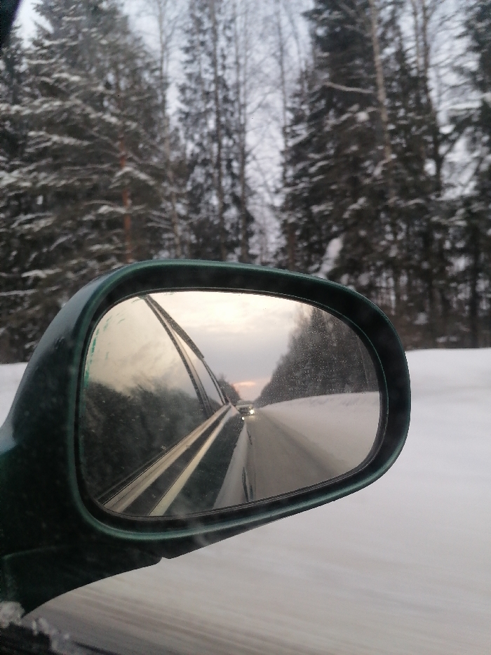 The image captures a winter scene from the perspective of inside a moving vehicle. The dominant feature is the car's passenger-side mirror, which occupies a significant portion of the foreground and midground on the left. The mirror's casing is a dark green color, slightly frosted or dirty with what appears to be snow and grime, indicating cold weather conditions.
Through the main section of the image, to the right of and above the mirror, a snowy forest is visible. Tall evergreen trees, heavily laden with snow on their branches, line the road. Some deciduous trees with bare branches are also scattered among the evergreens. The sky above the forest is overcast and light grey, suggesting a cloudy day, possibly late afternoon or early evening due to the soft, diffused light.
The mirror itself reflects the scene behind the car. A snow-covered road stretches into the distance, appearing as a wide, white expanse. In the far distance on the road, the bright headlights of another vehicle are visible, indicating a car following. The sky reflected in the mirror shows a similar overcast condition, but with a subtle hint of warmer, perhaps pinkish or yellowish, light near the horizon, reinforcing the possibility of late afternoon. Part of the car's own bodywork or interior is also reflected in the mirror's glass, creating a layered effect with horizontal and diagonal lines overlapping the view of the road behind. Snow or ice crystals are scattered across the surface of the mirror, further emphasizing the cold environment.
The snow on the ground outside the mirror is smooth and undisturbed, suggesting a recently snow-covered landscape or a less-trafficked road. The overall impression is one of a tranquil, snowy journey through a winter forest. There is no visible text in the image.