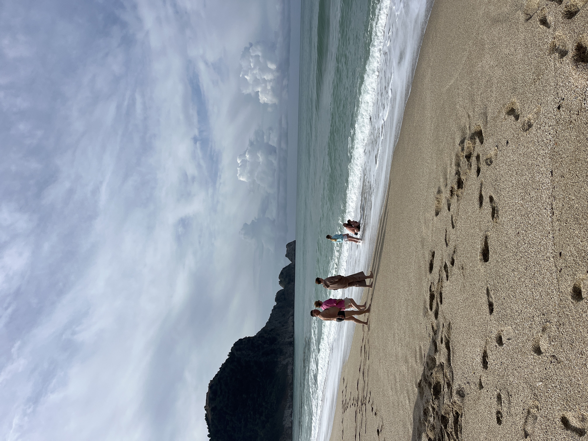The image depicts a beach scene, likely in Alanya, Turkey, considering the coastal features present. In the foreground, a sandy beach stretches out, marked with numerous footprints. Several people are walking along the shoreline where the waves meet the sand. One person is wearing a pink shirt, another a brown outfit, and a third is in black swim trunks. Further down the beach, other individuals are gathered closer to the water. To the left, a dark, mountainous headland rises sharply, indicating a coastal landscape characteristic of Alanya. The sky is overcast with various cloud formations, and the sea appears greenish with foamy waves breaking along the shore.