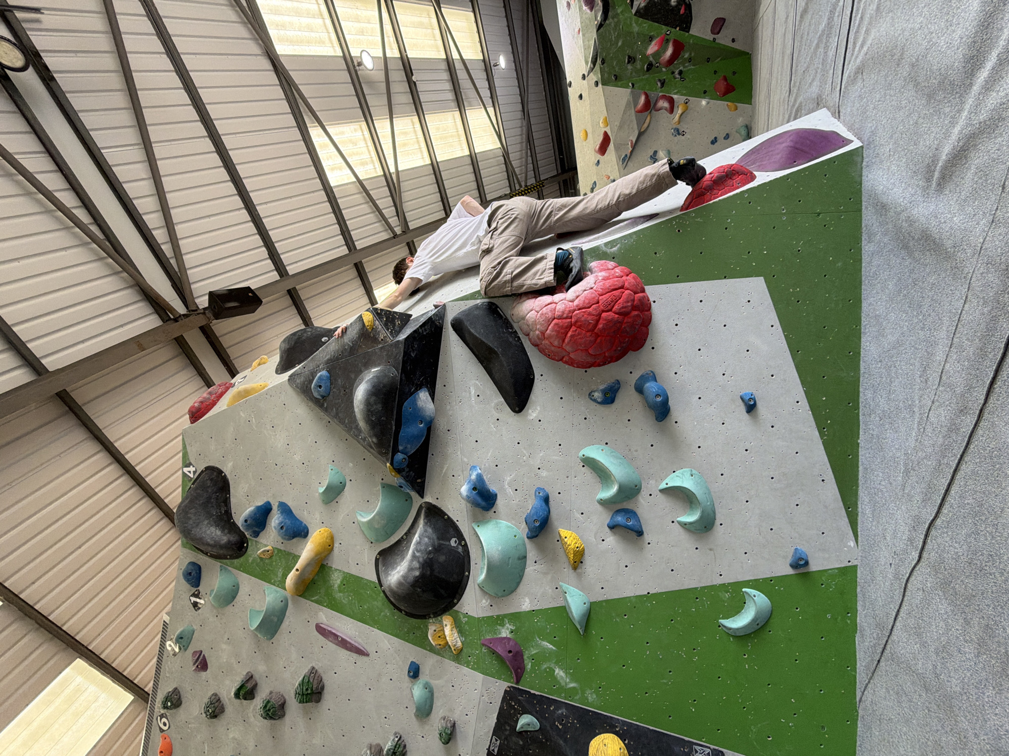 A man is bouldering indoors at a climbing gym. The artificial rock wall features various-shaped climbing holds in shades of blue, black, red, yellow, and purple. The man wears a white t-shirt and khaki pants. He is stretched out on the wall, with one foot resting on a red hold. The gym has a metal ceiling with industrial lighting. This is located at the Ipswich Boulder House in Ipswich, United Kingdom.