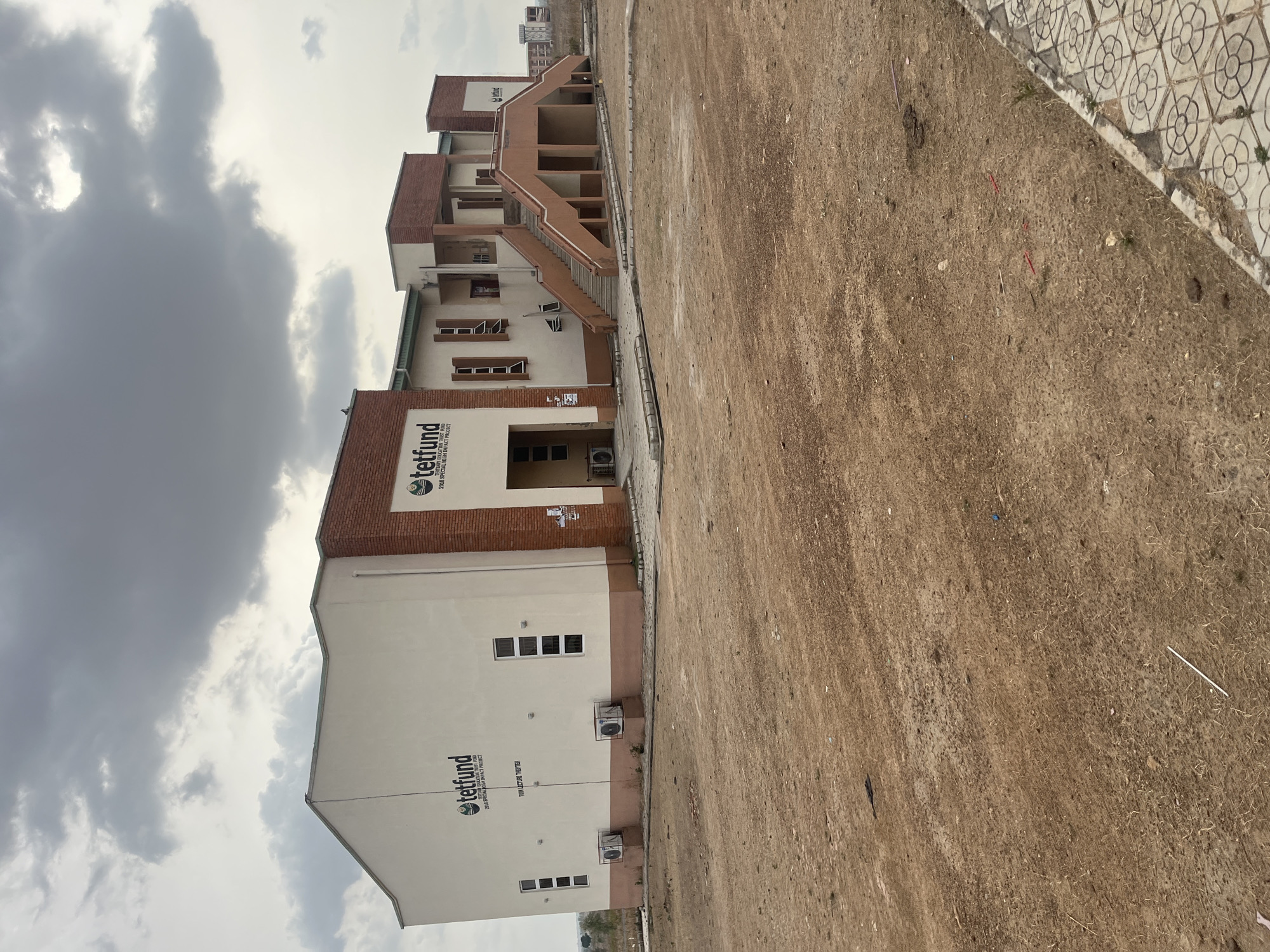 The image depicts a building in Baganakwo, Nigeria, with a sign indicating it's a TETFUND (Tertiary Education Trust Fund) project, specifically a "Twin Lecture Theater" from the 2018 Special High Impact Project. The building is multi-storied with a brick facade on the upper sections and light-colored walls. There are exterior staircases connecting different levels. Air conditioning units are visible on the side of the building. The foreground shows a dirt or gravel-covered ground area with some debris. The sky is overcast with heavy clouds.