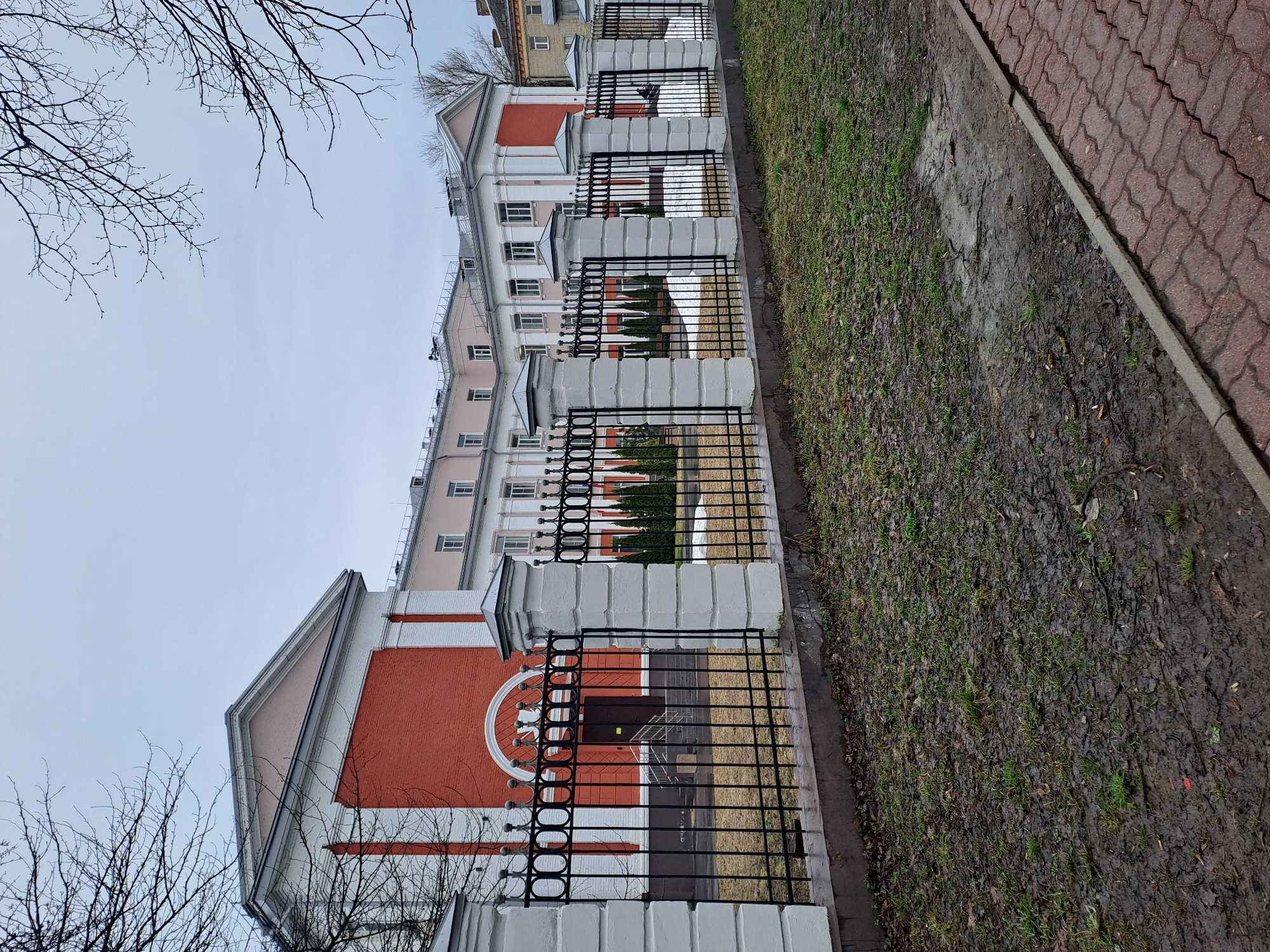 The image shows a building in Elektrostal, Russia. It's a multi-story structure with a red and white facade. A black metal fence runs along the front of the building. The ground on the side of the fence consists of muddy grass. A sidewalk made of red pavers is visible on the right side of the image. Bare tree branches are present in the top left corner. The sky is overcast.