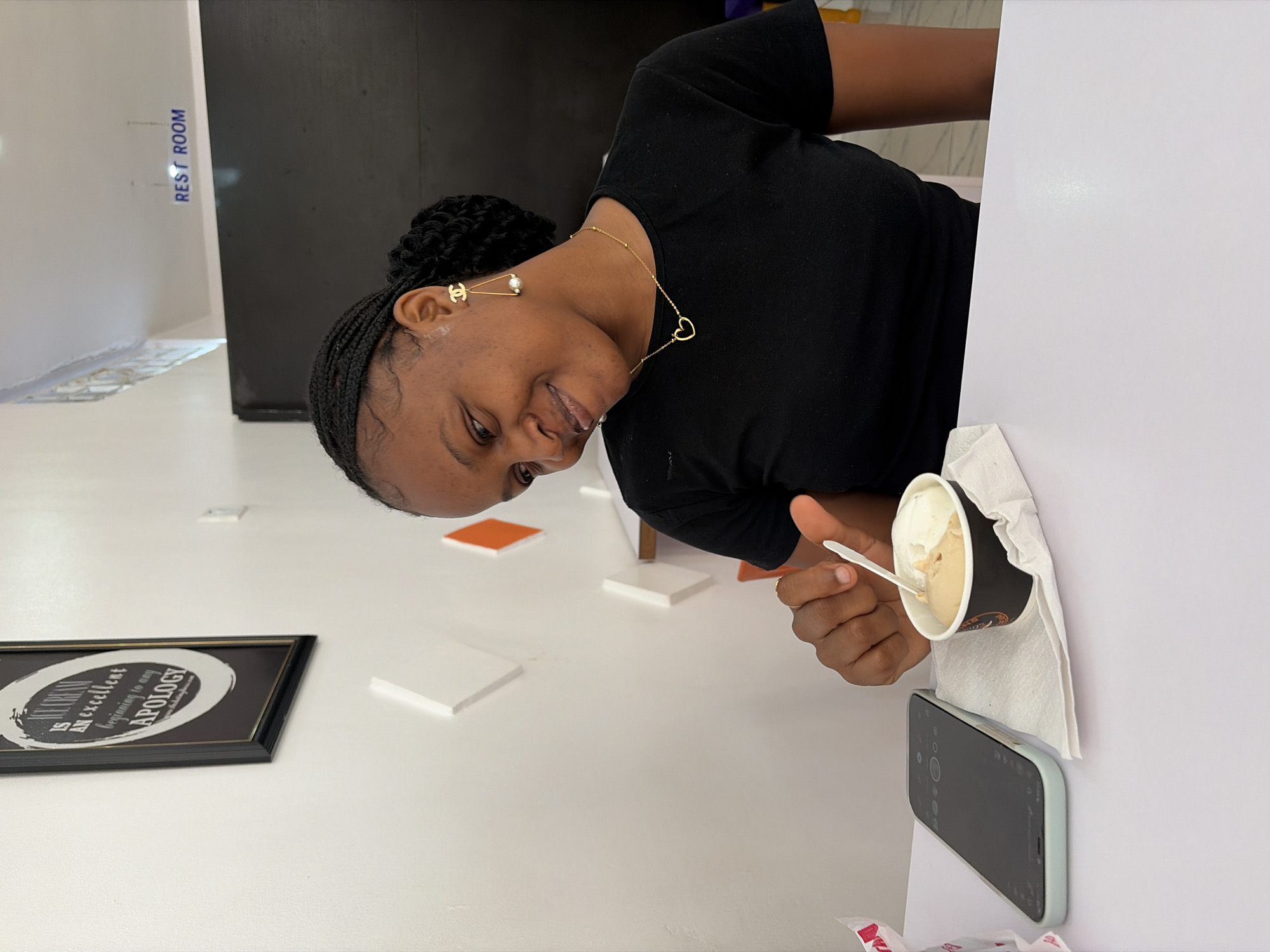 The image captures a smiling woman indoors, likely in a cafe or ice cream establishment. Her upper body is visible, and she looks towards the viewer with a cheerful expression. She has dark skin and braided hair pulled back into a bun, wearing a black short-sleeved top. Her accessories include a delicate gold necklace with a heart pendant and gold earrings; one features an interlocking 'CC' logo (Chanel) with a pearl, and the other is a pearl drop with a geometric shape.
In her right hand, she holds a paper cup of ice cream with a white plastic spoon. The cup contains two scoops, appearing to be vanilla and a lighter brown flavor, and rests on a white napkin. Adjacent to the ice cream, a smartphone with a light green case lies flat on a white surface.
The setting features bright, even lighting, characteristic of an indoor environment. A white wall in the background prominently displays the blue text "REST ROOM." On a white surface in the foreground, a black-framed picture is visible, with partially obscured white text reading, "...TRUTH IS AN excellent beginning to any APOLOGY." Small white and orange square objects are scattered across the white surface. The background also includes a dark wall or partition. The woman's expression conveys happiness and relaxation.