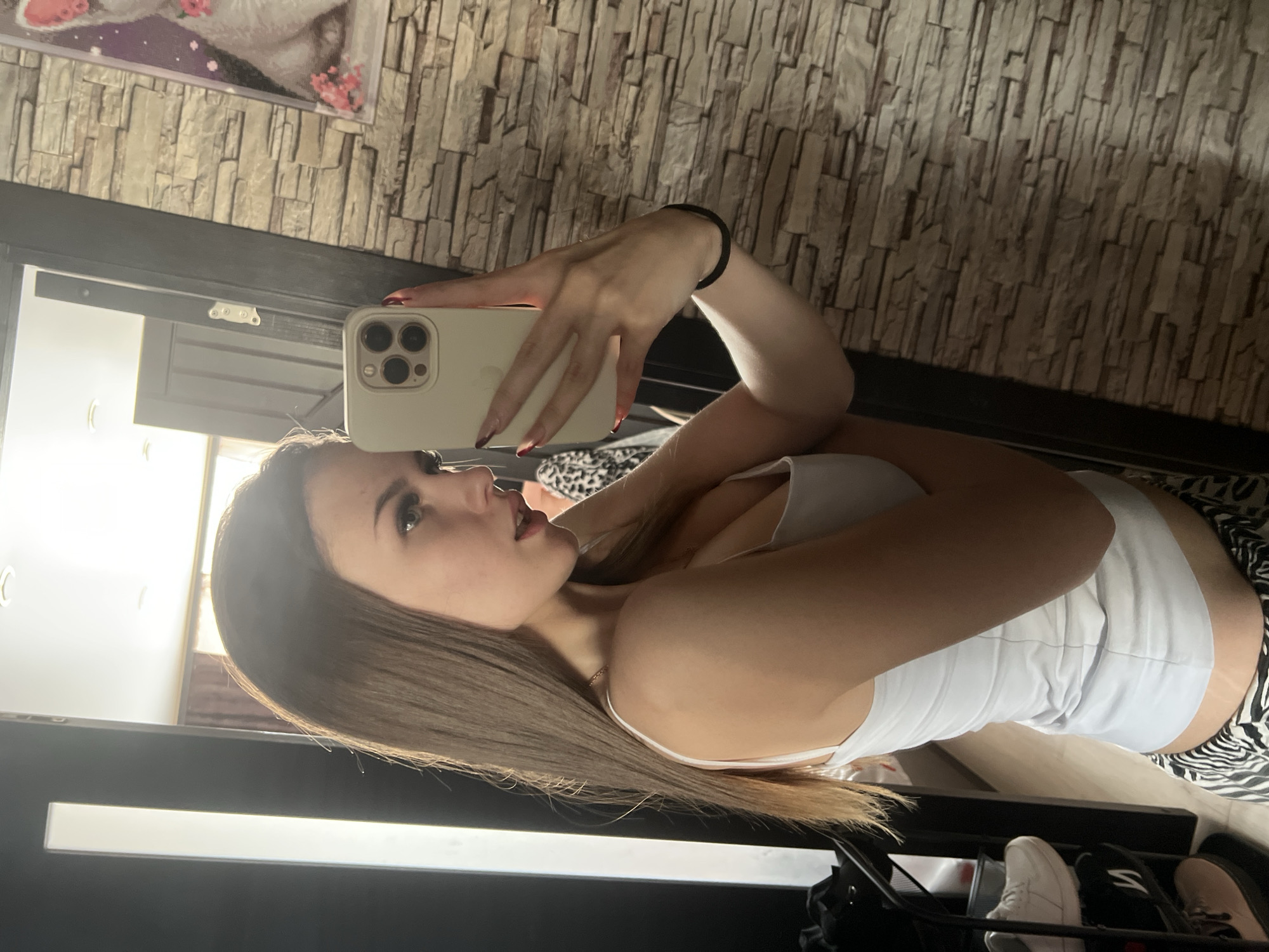 A young woman with long, straight brown hair is taking a selfie in a room that appears to be her bedroom. She is wearing a white tank top. She holds a smartphone up with her right hand, which has painted fingernails. She is looking at the phone's screen. In the background, there is a stone-patterned wall and a framed picture above a doorway. In the foreground, a black item, possibly furniture or a rack, and a pair of white shoes are visible. It is not possible to confirm the location in Noviy, Russia from the provided image.