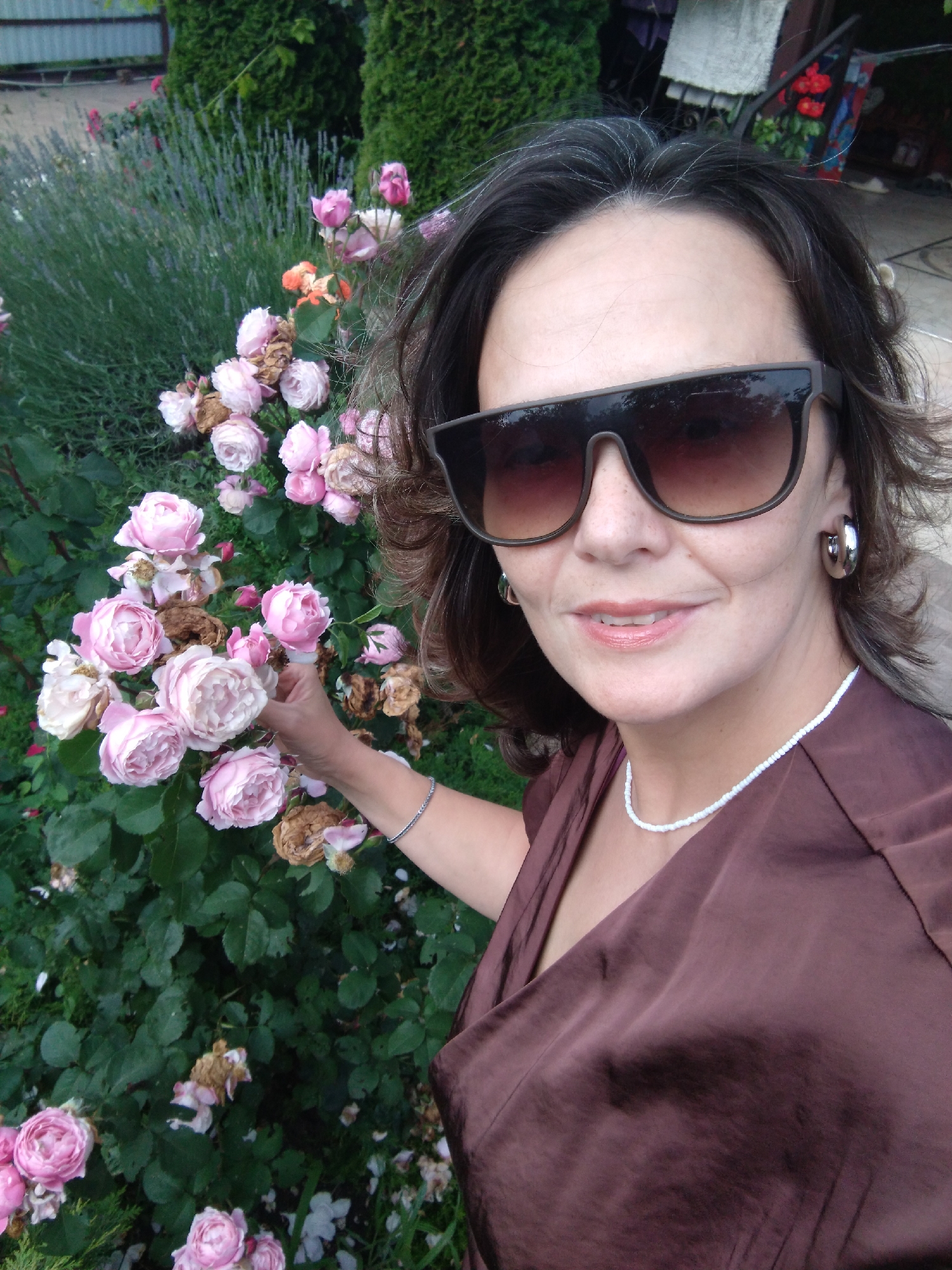 A medium shot captures a woman standing outdoors in what appears to be a garden in Vinogradny, Russia. The woman, facing the camera and smiling, wears oversized dark sunglasses, large silver hoop earrings, and a brown satin dress. A delicate white beaded necklace is visible around her neck. Her dark hair, with streaks of grey, is partially obscured by her sunglasses and hair. Her left hand is extended and touching a cluster of pink roses, some of which are wilted. Behind her, a dense bush of roses and lavender can be seen, along with greenery and what appears to be a fence in the background. The lighting suggests it is daytime.