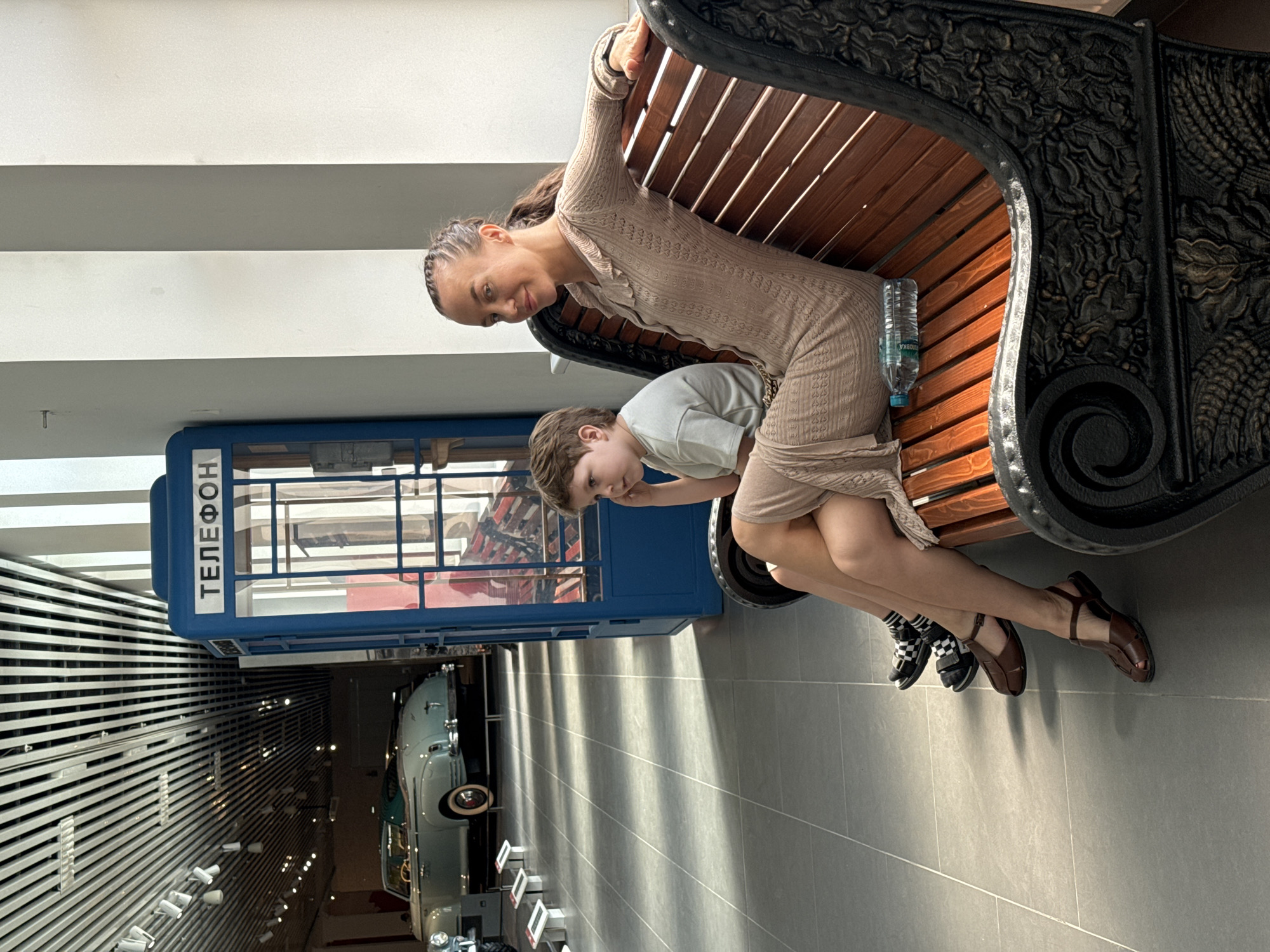 A woman and a child sit on a wooden and iron bench next to a blue telephone booth inside the Military Museum in Verkhnyaya Pyshma, Russia. The woman wears a tan dress and brown sandals. The child wears a gray t-shirt and black and white checkered shoes. A water bottle sits on the bench next to the woman. Beyond the telephone booth, a vintage car is visible. The museum interior features gray floors and a slatted ceiling.