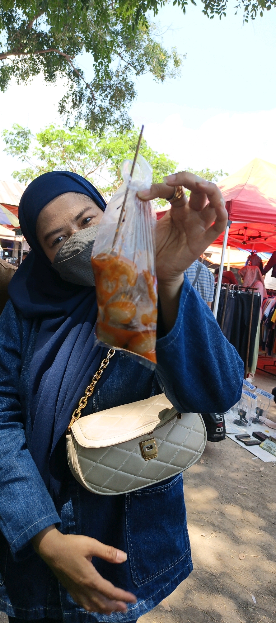 A woman wearing a blue hijab and denim jacket is holding a clear plastic bag with food items in an outdoor market. The bag is skewered with a wooden stick and contains what appear to be meatballs or dumplings in an orange sauce. The woman is wearing a grey face mask, and a quilted, cream-colored handbag with a gold chain strap is slung across her body. Behind her, red and yellow tents are visible, as well as racks of clothing. The ground is covered in dirt and scattered leaves. The setting appears to be a bustling street market, possibly in Jatinom, Indonesia.