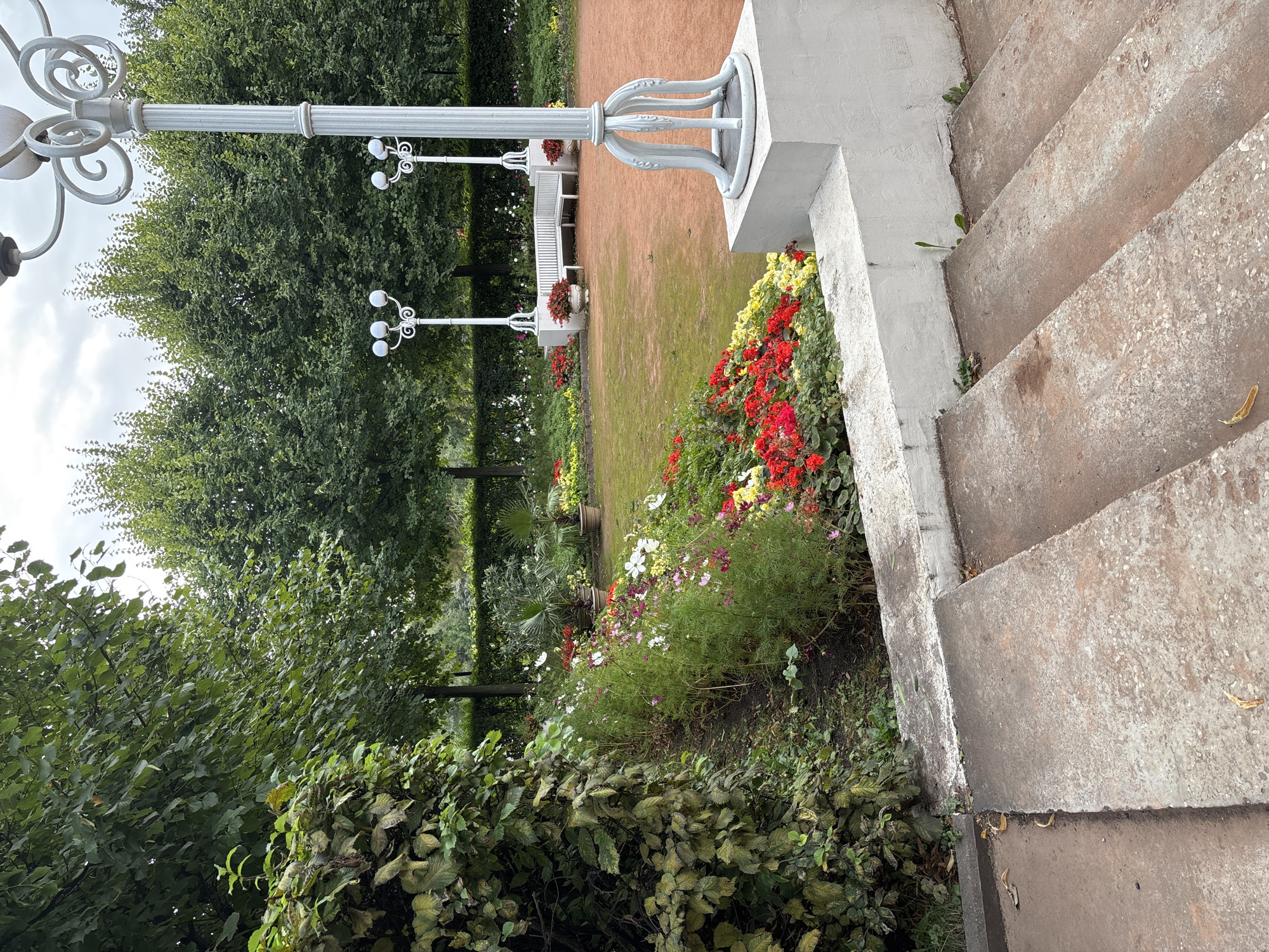 The image depicts a formal garden with a central path, flowerbeds, and decorative lampposts. In the foreground, a set of concrete steps leads up to a raised area. To the left, a dense hedge is visible, and beyond it, a sloping flowerbed is filled with an assortment of red, white, and pink flowers. The path is made of reddish-brown gravel, and in the background, a white bench is situated in front of a row of trees. Two ornate lampposts with multiple globes stand on either side of the path, adding to the garden's aesthetic. The sky is overcast, suggesting a cloudy day. The scene appears to be in a park or botanical garden, possibly in Moscow, Russia, given the presence of such manicured and ornate landscaping. There are no people or animals visible in the image.
