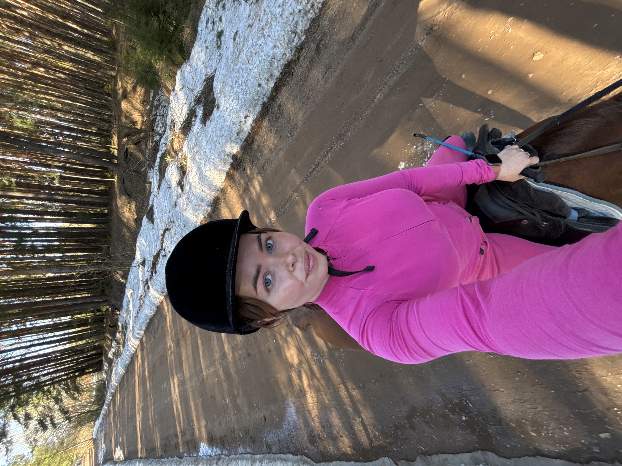A young woman is depicted horseback riding on a dirt path in a rural, forested area, likely in Русский Вожой, Russia. She is the primary subject, looking directly at the camera with a calm, slightly smiling expression. She has light skin, blue eyes, and light brown hair partially visible under a black equestrian helmet. Her attire consists of a bright pink long-sleeved top and matching pink riding pants. An Apple Watch is visible on her left wrist, and she holds the reins and a light blue riding crop. The brown horse she is riding is partially visible, showing its back, saddle, and parts of its neck.
The setting features a wide dirt path, bordered on one side by patches of melting snow, suggesting early spring conditions. A dense forest of tall, slender evergreen trees lines the path. Strong sunlight casts long, distinct shadows from the trees across the path, indicating either early morning or late afternoon. The image is taken from a slightly high, tilted angle, giving a selfie-like perspective. No visible text is present in the image.