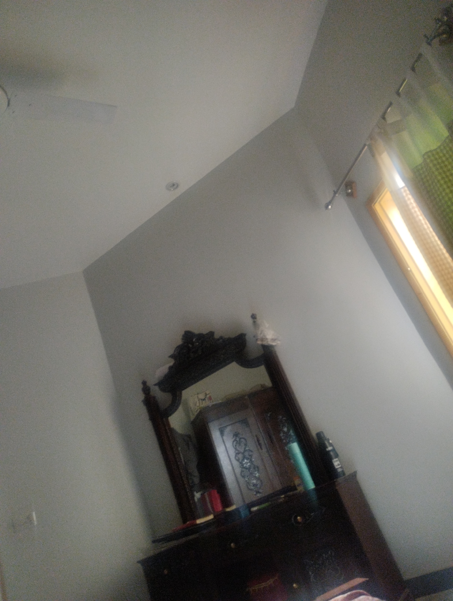 The image shows an interior view of a room, likely a bedroom, with a dresser and mirror. The dresser is dark brown wood, with several drawers visible. On top of the dresser, there are a few items, including what appears to be a teal-colored bottle, a dark-colored bottle, and a red object. The mirror above the dresser has an ornate, dark wooden frame. Reflected in the mirror is a wooden cabinet with decorative detailing on its doors. The walls are painted a light gray color, and the ceiling is also light-colored. A window with yellow and green curtains is visible on the right side of the image, suggesting daylight. There are no people or animals present in the image. The overall impression is that of a domestic setting.