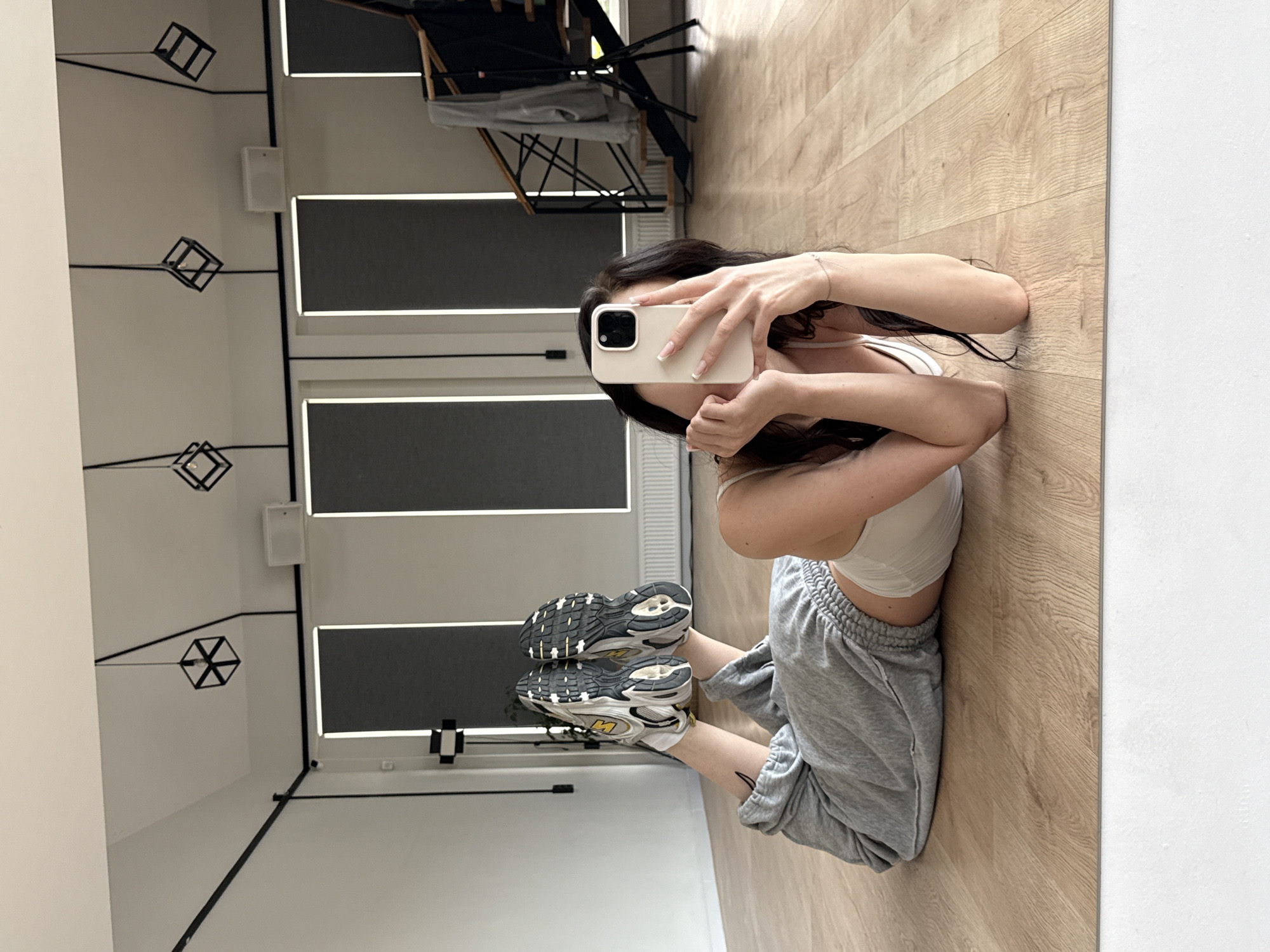 A full-body mirror selfie shows a young woman lying on her stomach on a polished wooden floor in what appears to be a dance studio or gym. She is wearing light grey sweatpants and a white sports bra, with athletic shoes on her feet. Her hair is dark and flows around her. She holds a light-colored smartphone in her right hand, obscuring her face as she takes the photo. In the background, the studio features large, dark-grey roller blinds covering windows, modern geometric pendant lights hanging from the ceiling, and a black staircase with railings. The overall environment is clean and modern. The location is identified as Agalarov Estate, Russia.