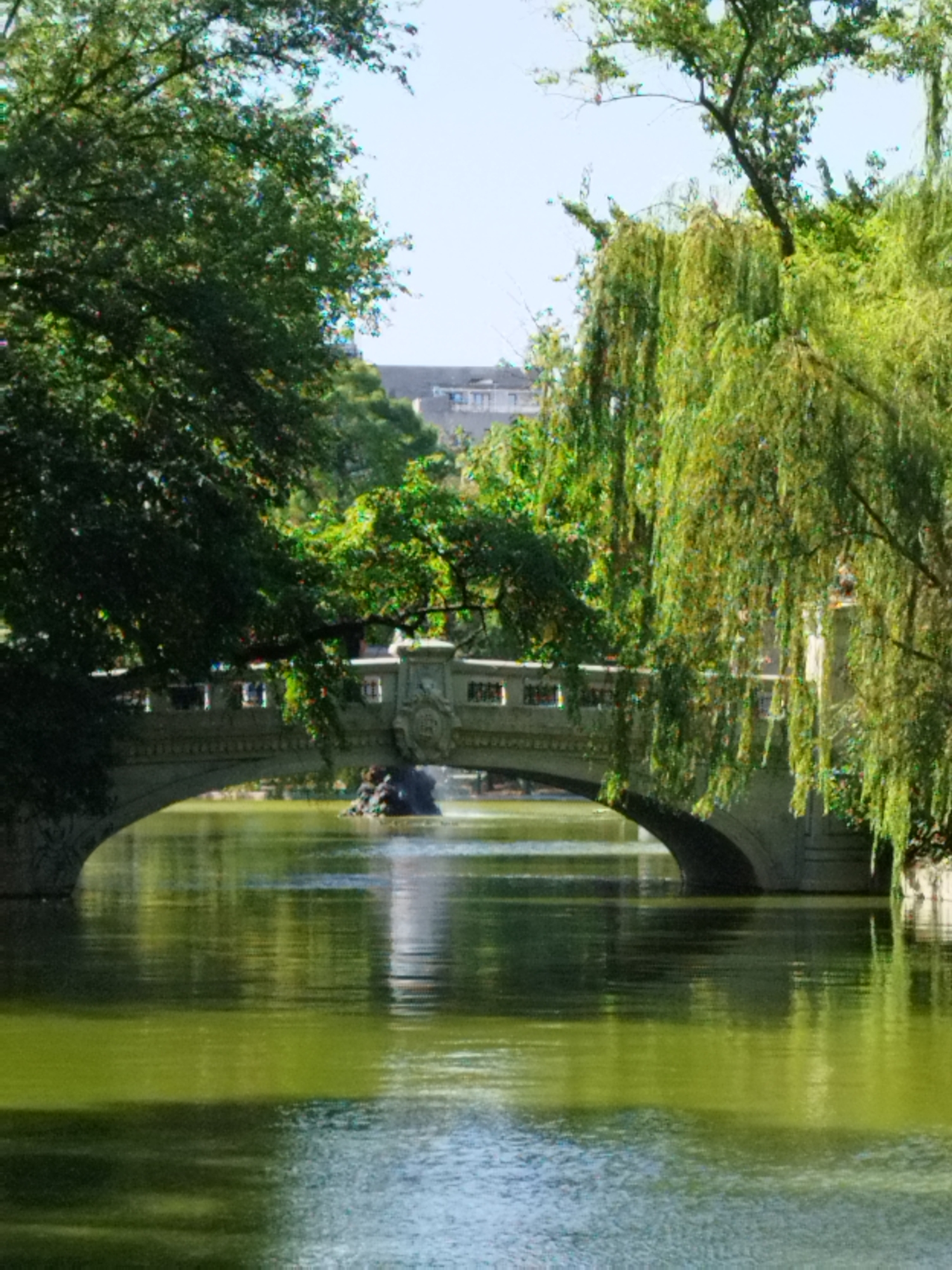 The image captures a serene water scene with a stone bridge and lush greenery, likely located in Bucharest, Romania. The bridge, constructed of stone, features an arched structure that spans across a tranquil body of water. The water's surface is a reflective green, mirroring the abundant foliage that surrounds it. On the right side of the image, weeping willow trees with their characteristic drooping branches add a touch of elegance to the landscape. The overall atmosphere is peaceful and picturesque, evoking a sense of calm and natural beauty within an urban setting. While the specific location within Bucharest is not identified, the presence of a bridge over water and extensive greenery suggests a park or a similar recreational area.