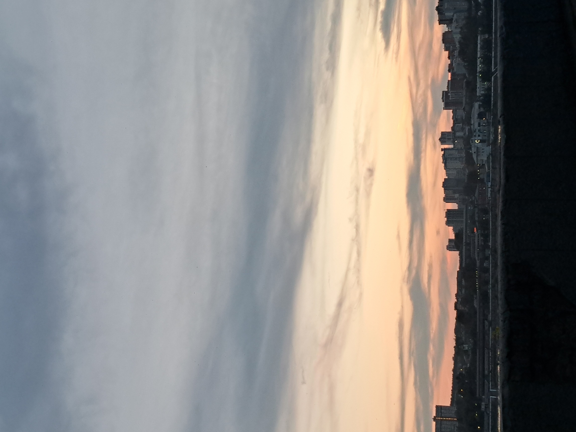 The image depicts a cityscape at dusk, likely from a vantage point overlooking the city of Irkutsk, Russia. The sky is filled with a gradient of colors, transitioning from a pale orange near the horizon to a muted gray higher up, with streaks of clouds creating texture. The lower portion of the image features the silhouette of the city skyline, with numerous buildings of varying heights, including apartment blocks and other structures. In the foreground, a dark, textured surface, possibly a wall or embankment, obscures the very bottom of the frame. The overall impression is one of a quiet evening over an urban landscape.