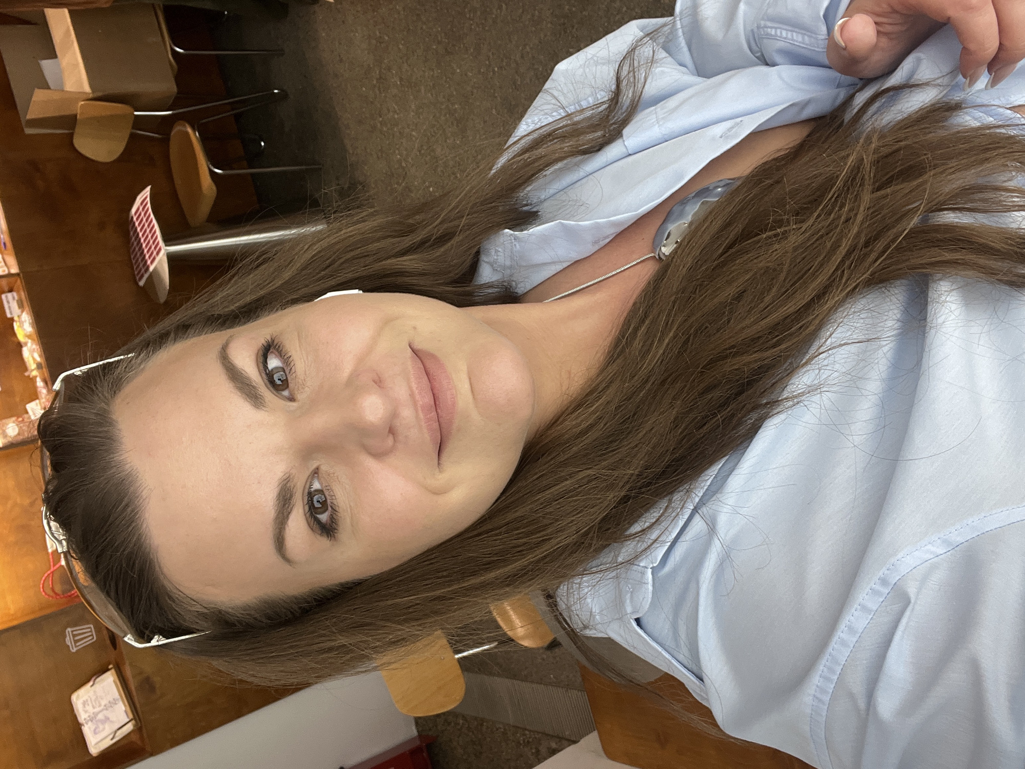A selfie taken by a Caucasian woman with brown hair wearing a light blue button-down shirt and a necklace, likely indoors based on the background with chairs and shelving. It is impossible to confirm the location is Novosibirsk, Russia, or to provide a forensic description without additional information.