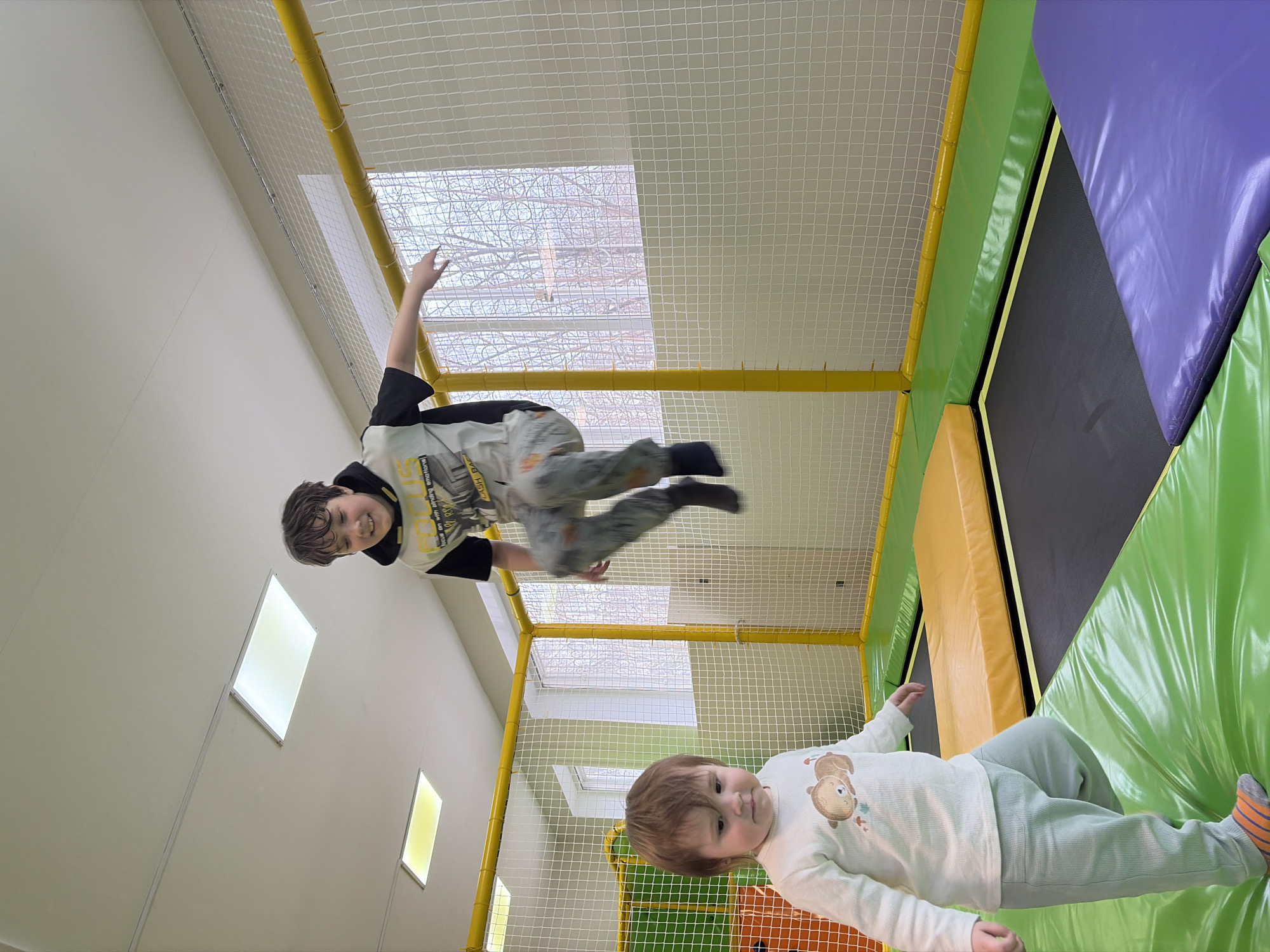 Two children are playing on a trampoline inside an indoor play area. The older child is airborne, suspended in mid-jump near the top of the frame, with arms outstretched and legs bent. They are wearing a black, white, and gray patterned shirt. The younger child is lying on a padded area, wearing a white shirt with a bear graphic and light green pants. The trampoline is surrounded by a safety net with yellow supports and various colored padded surfaces: purple, green, and yellow. The walls are light-colored with rectangular light fixtures. Through the ceiling netting, bare tree branches are visible, suggesting an outdoor view from the play area. The location is implied to be Chaikovsky, Russia based on the prompt.